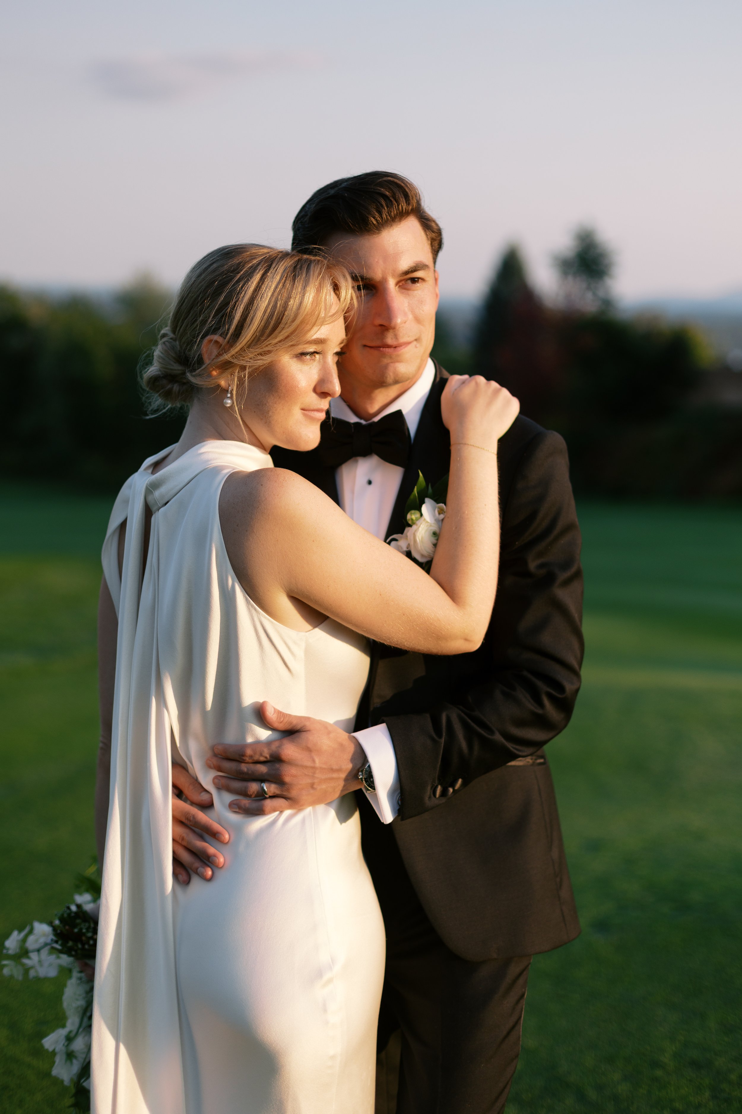 A bride and groom embrace outdoors on their wedding day, with the bride in a white dress and the groom in a black tuxedo, smiling and looking into the distance during golden hour.