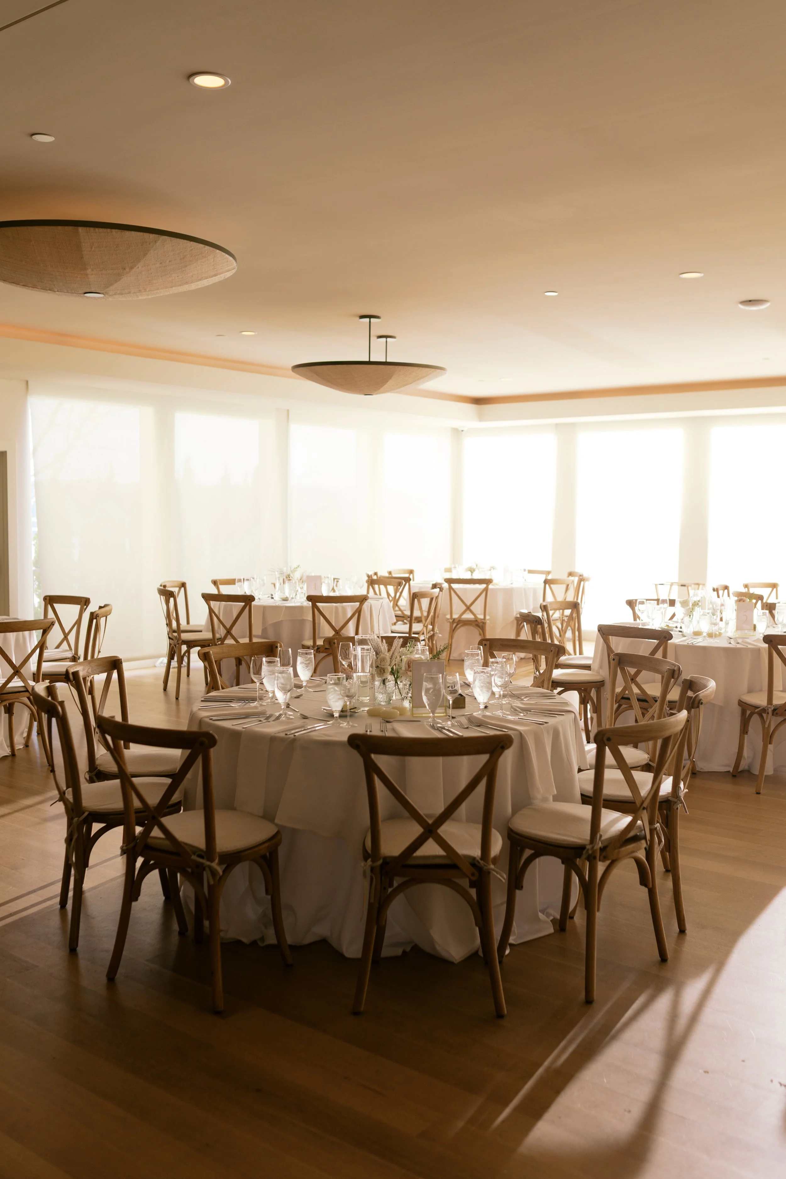 Elegant banquet room with round tables covered in white tablecloths, set with glassware and floral centerpieces, illuminated by natural light from large windows.