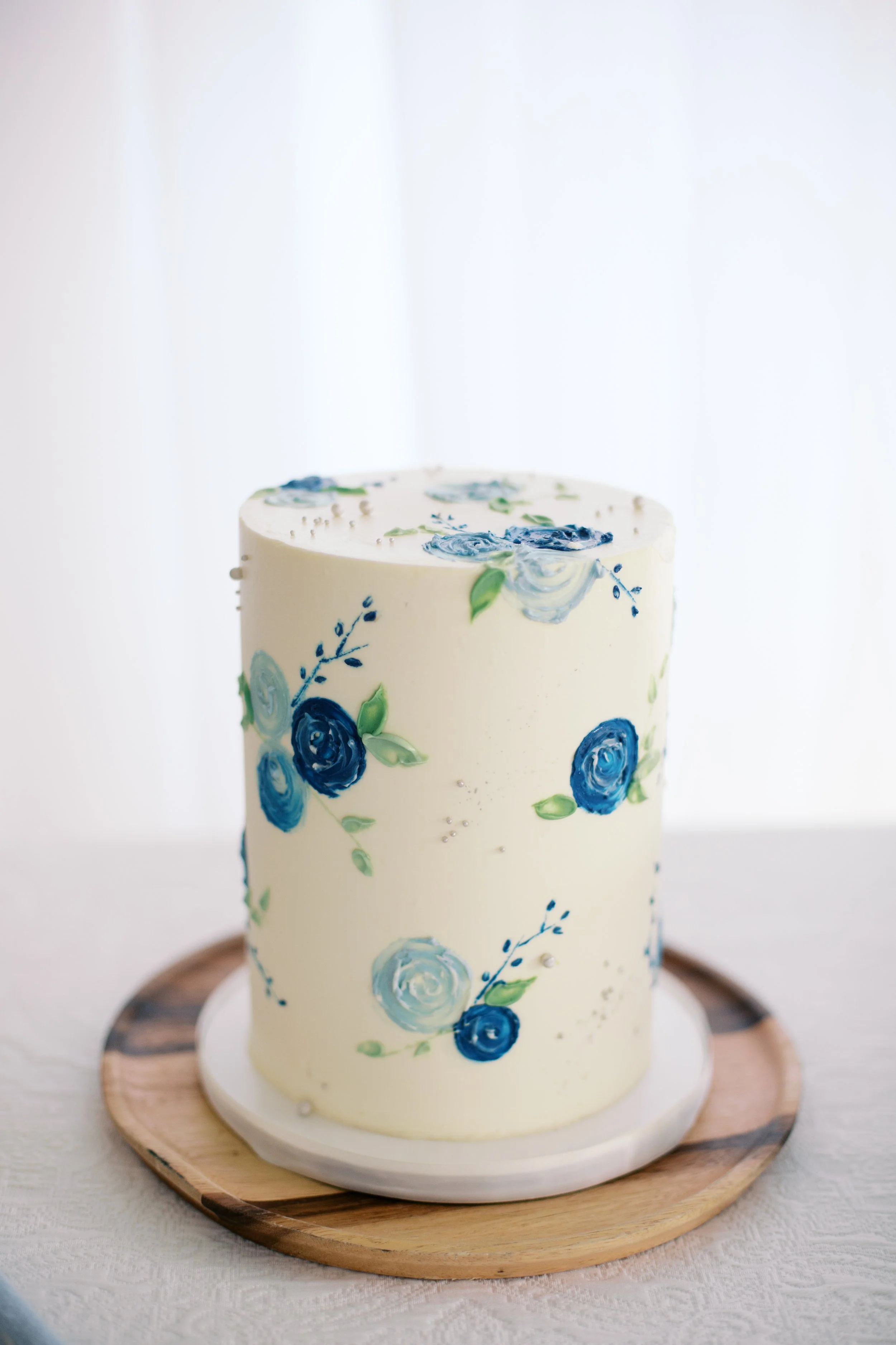 A tall, cylindrical white cake decorated with blue and green flower designs on a wooden cake board.