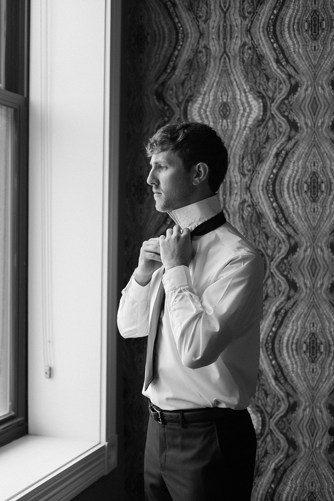 A man is standing by a window, adjusting his black bow tie while looking outside. He is wearing a white dress shirt, dark pants, and a black belt, with a patterned wall in the background.