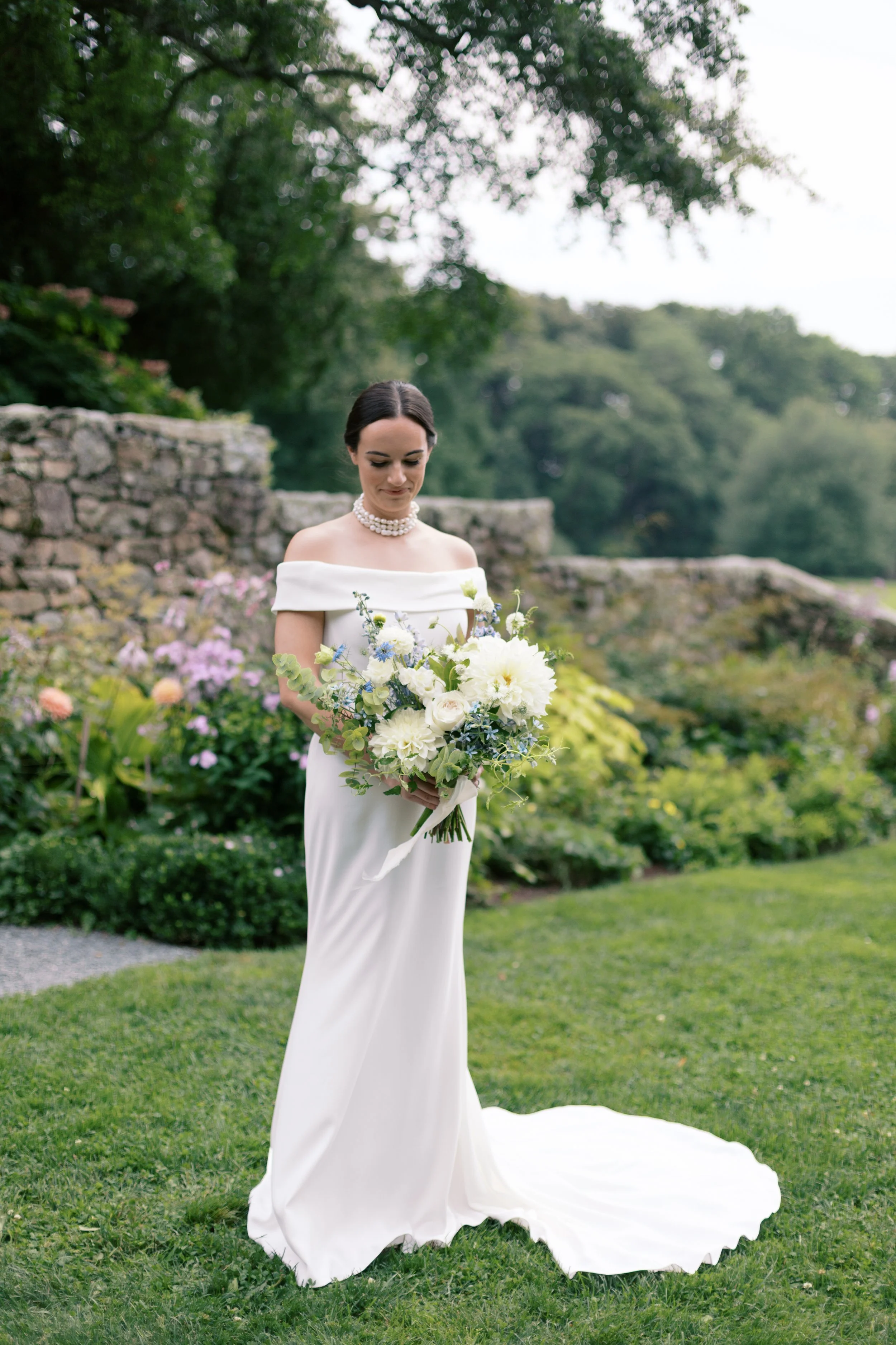 A bride standing on a grassy lawn, holding a bouquet of white and blue flowers, wearing a white off-the-shoulder wedding gown and pearl necklace, with a stone wall and garden in the background.