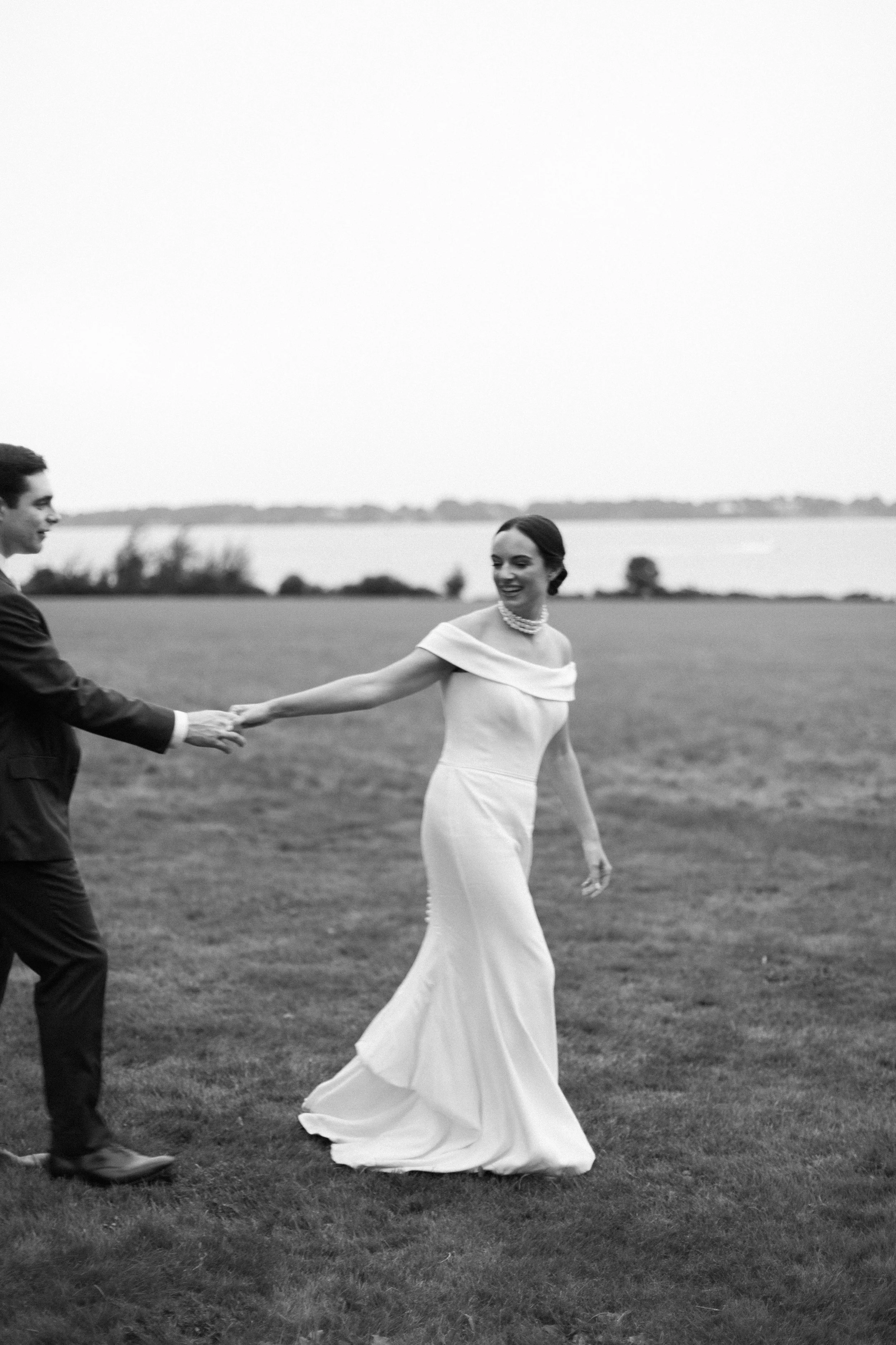 A black and white photo of a woman in a long, elegant dress holding hands with a man in a suit on a grassy field near a body of water, smiling and looking at each other.