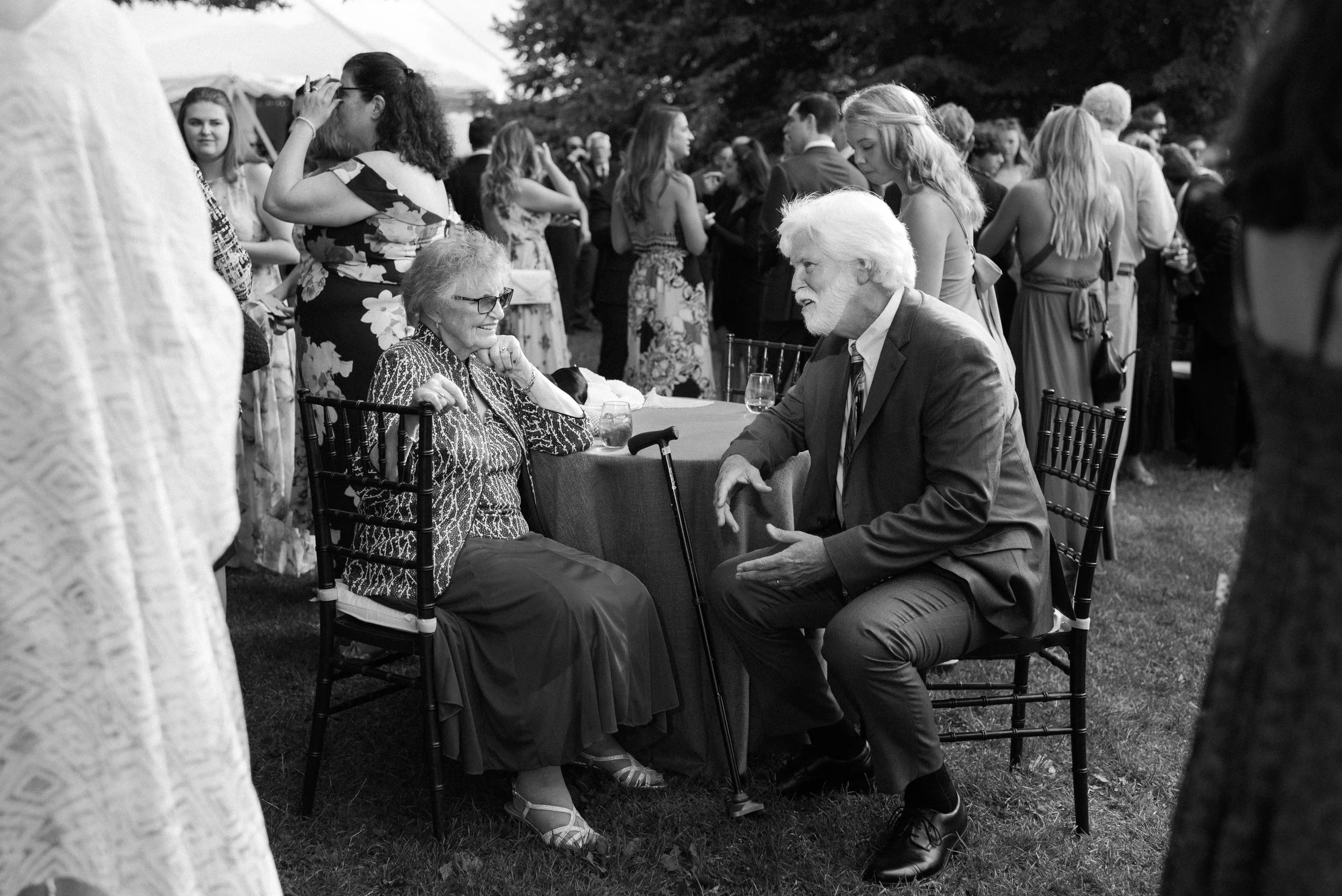 An elderly woman and man engaging in a conversation at a social outdoor event, with many people in the background.