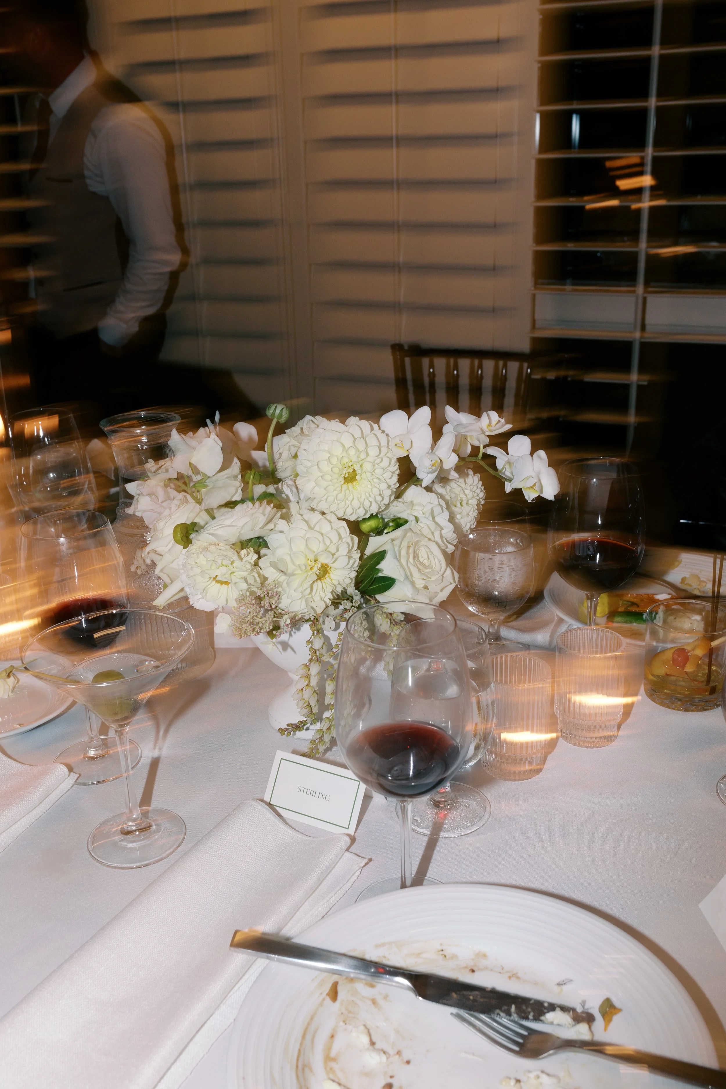 A table with a centerpiece of white flowers, several glasses of red wine, and candlelight, set for a formal dinner.