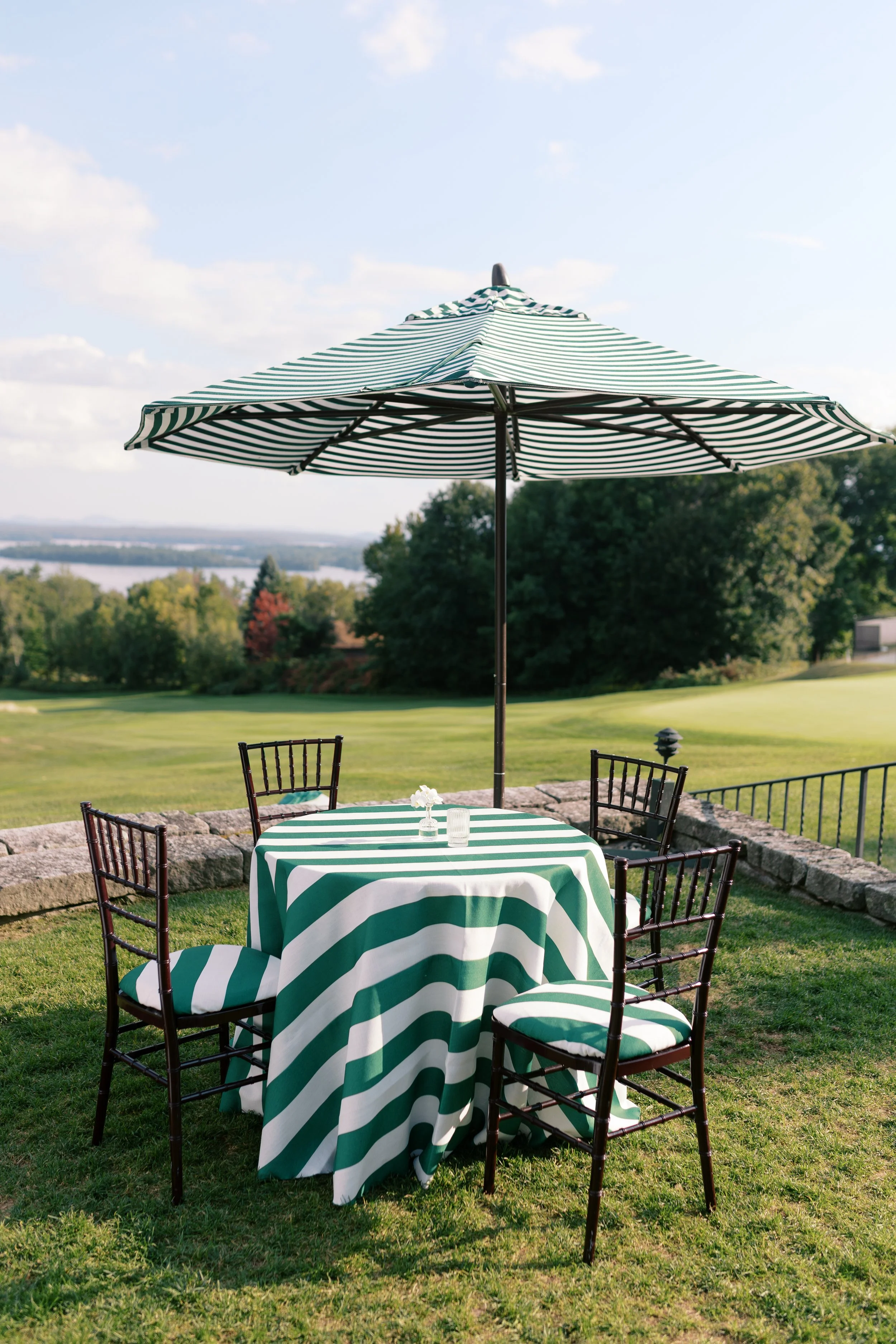Outdoor dining table with a green and white striped tablecloth, surrounded by four black chairs, under a green and white striped umbrella, on a grassy area with trees and a lake in the background.