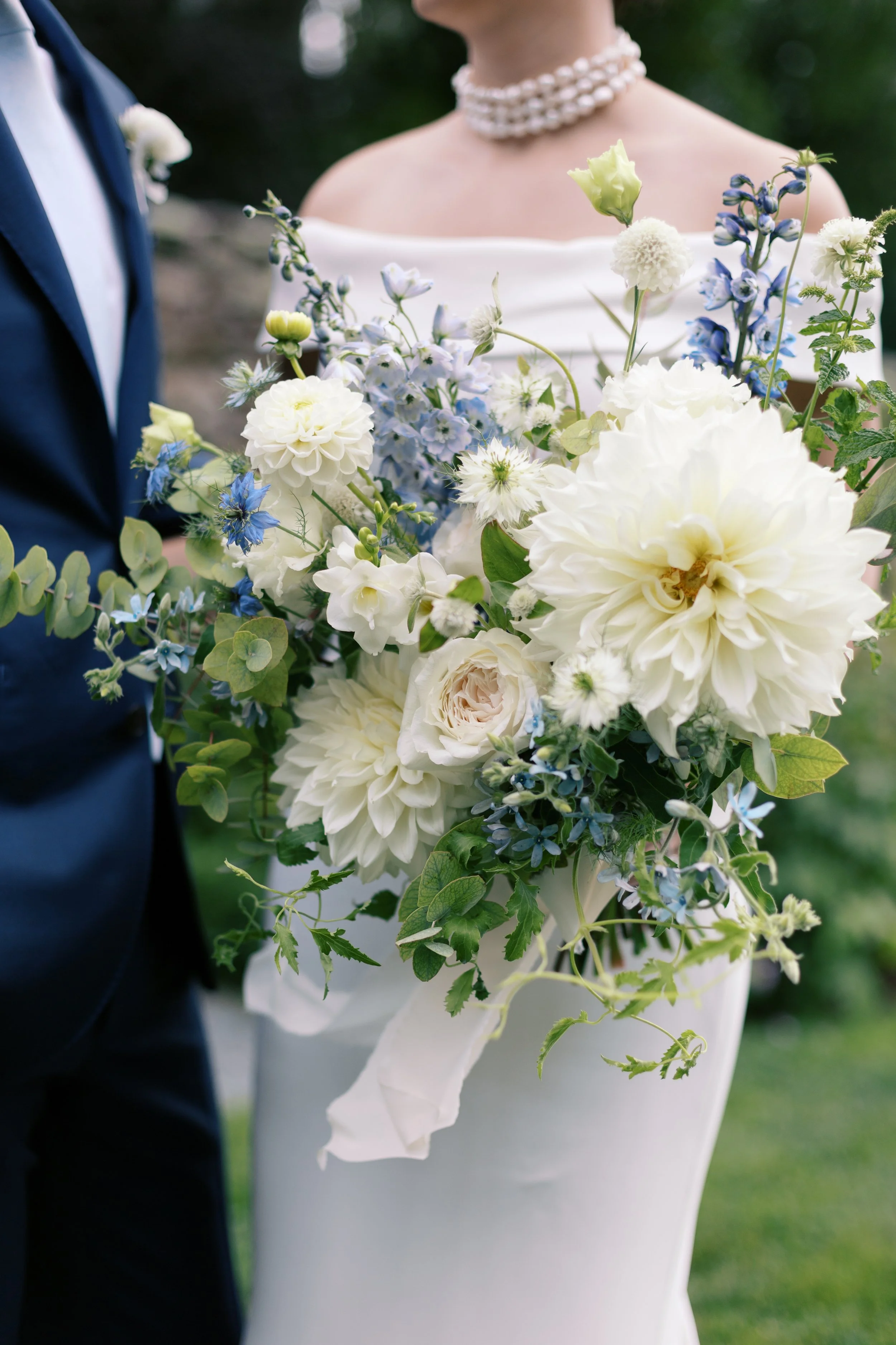 A bride holding a large bouquet of white and blue flowers at a wedding, with a groom partially visible beside her.
