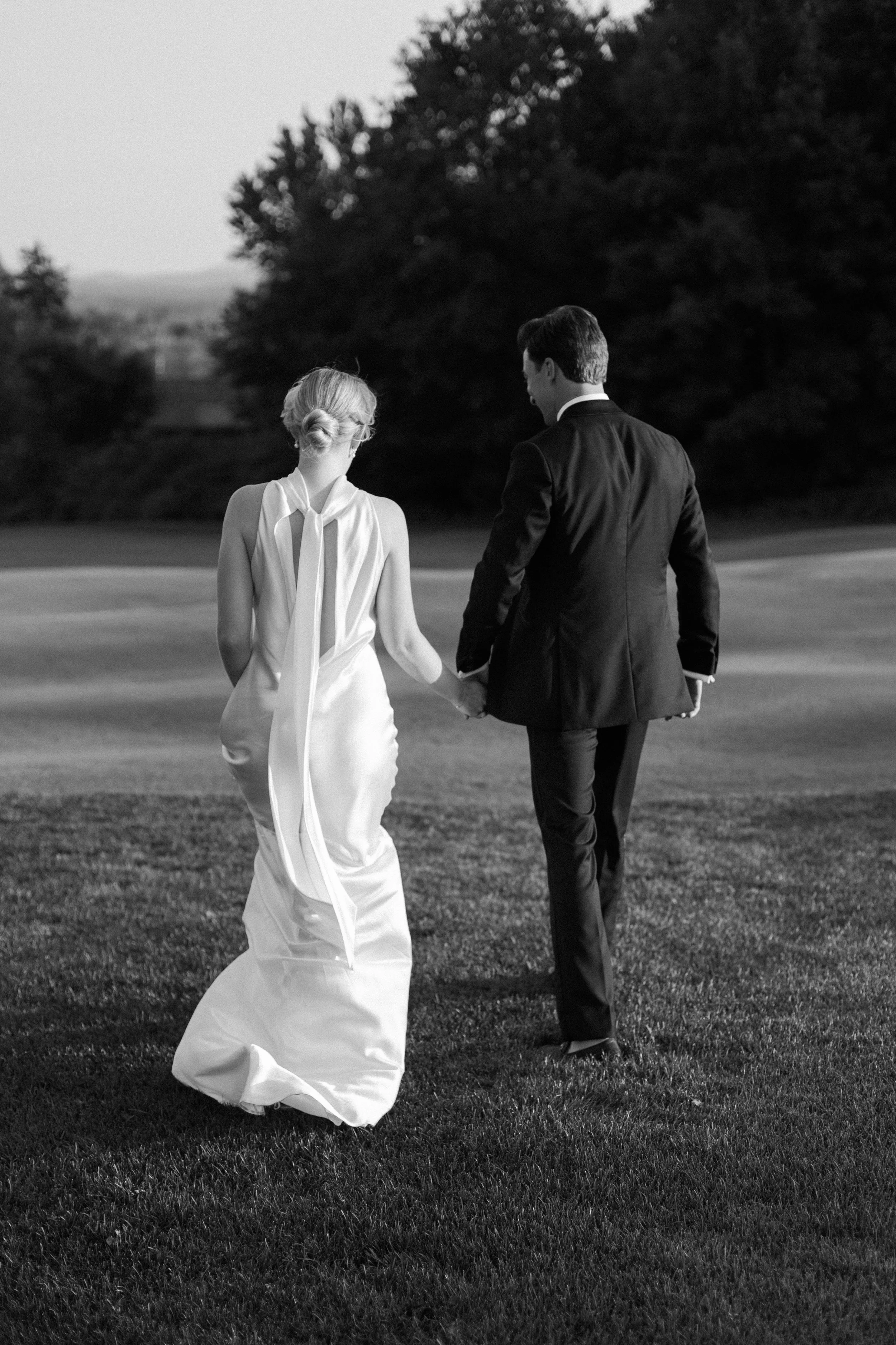 A black and white photograph of a bride and groom walking hand in hand across a grassy field, with trees in the background.