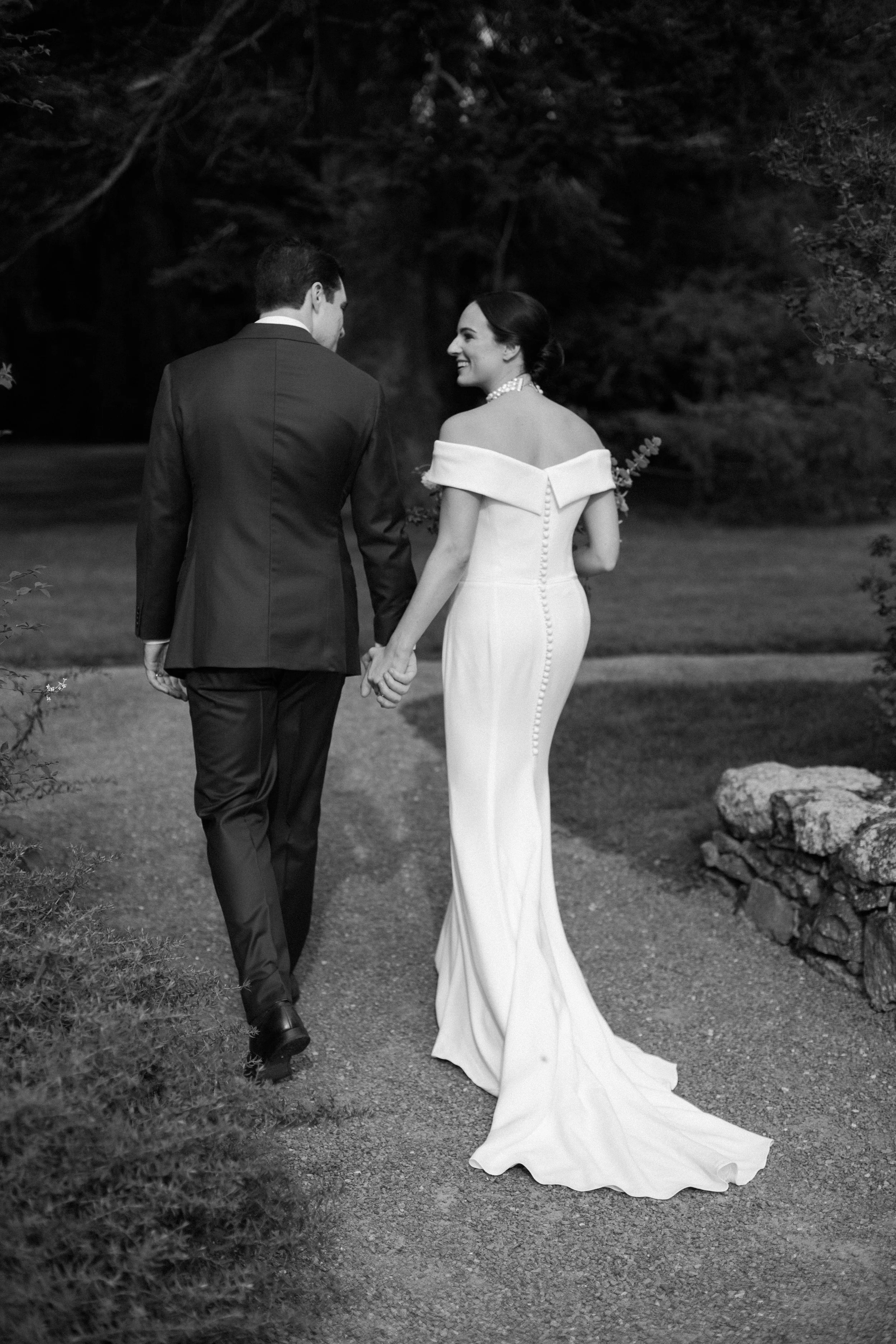 A black-and-white photograph of a bride and groom holding hands, walking outdoors on a gravel path surrounded by trees and rocks.