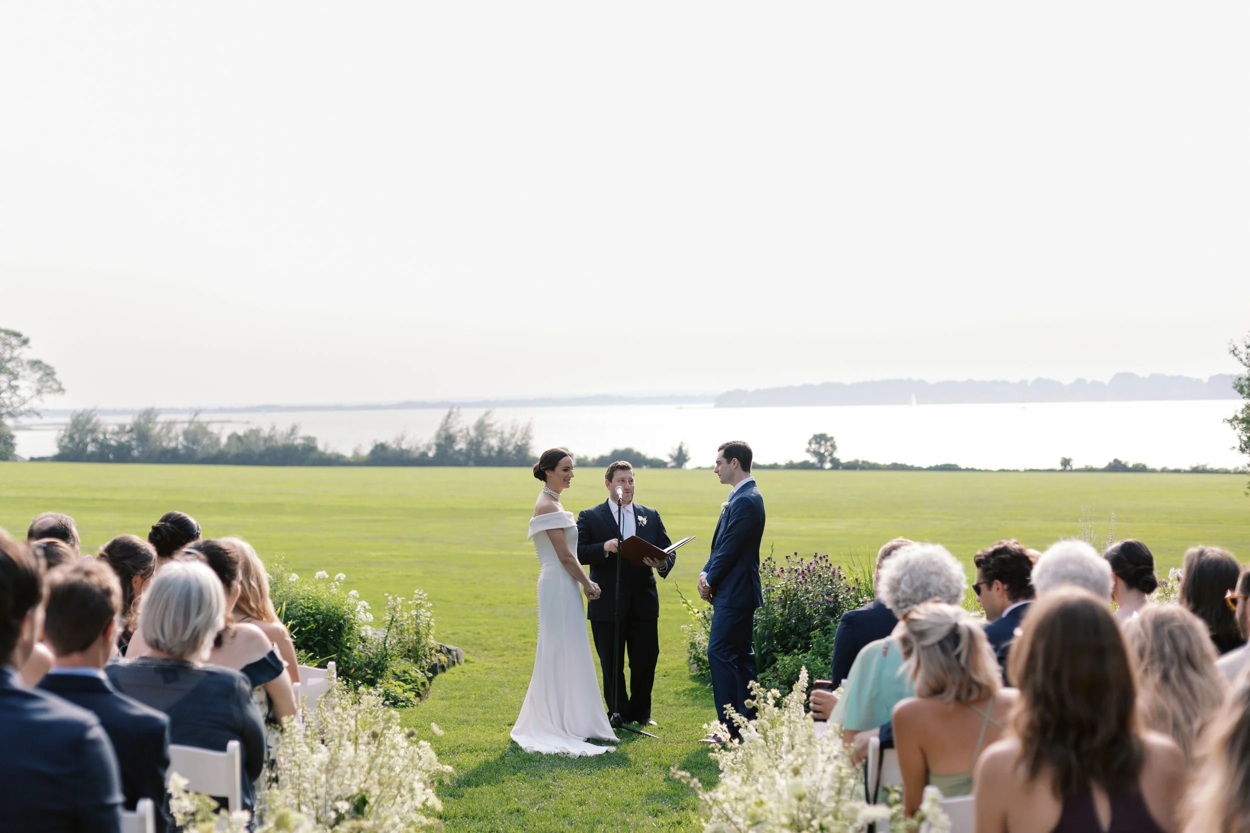 A wedding ceremony taking place outdoors on a green lawn by a large body of water, with the bride and groom standing before a officiant, surrounded by seated guests.