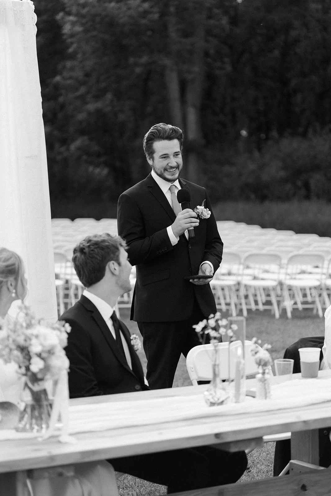 A man giving a speech at an outdoor wedding reception, dressed in a suit with a boutonniere, holding a microphone, with seated guests and decorated tables visible.