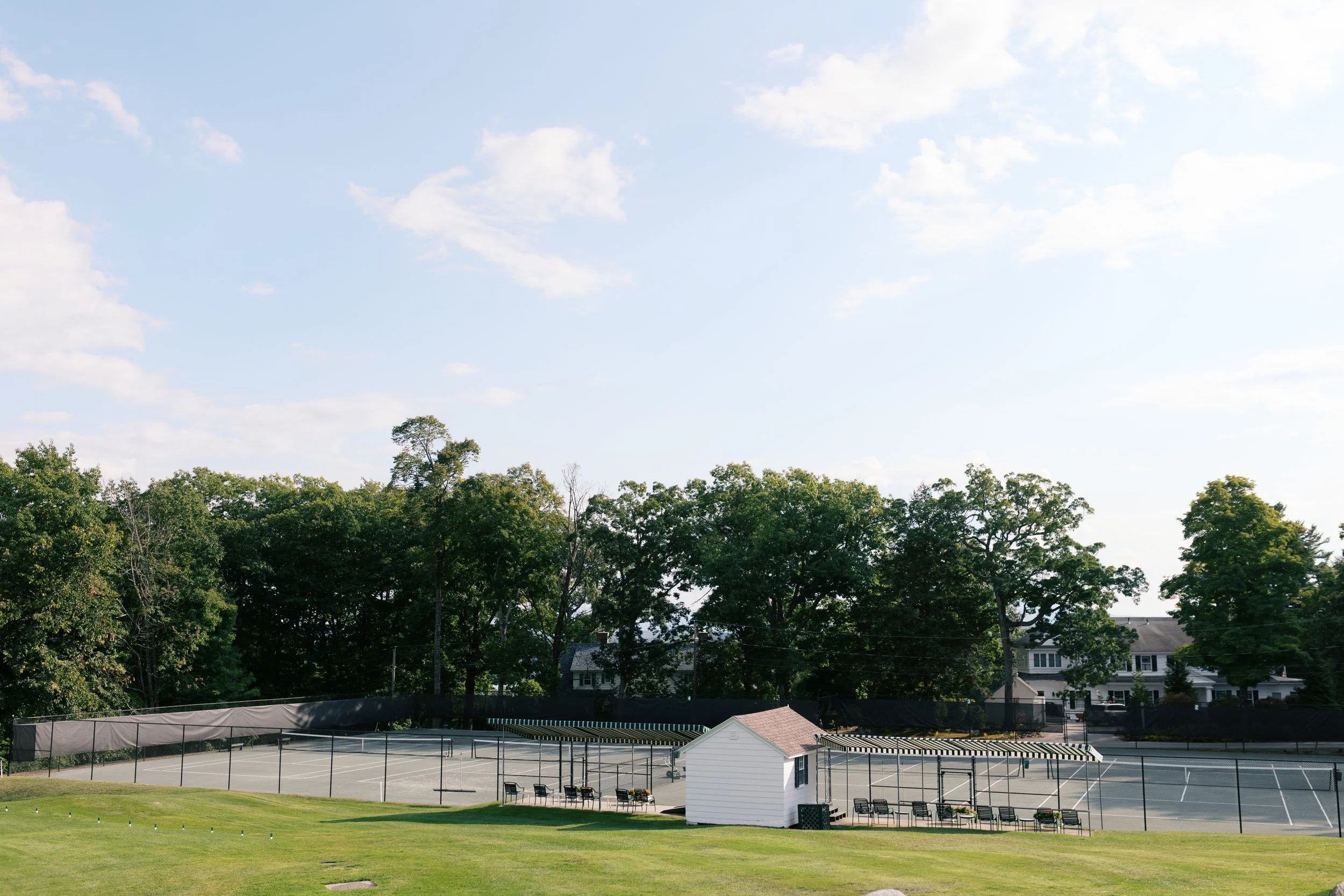 Empty tennis courts with a small white building, surrounded by black fencing, under a blue sky with scattered clouds, and houses in the background.