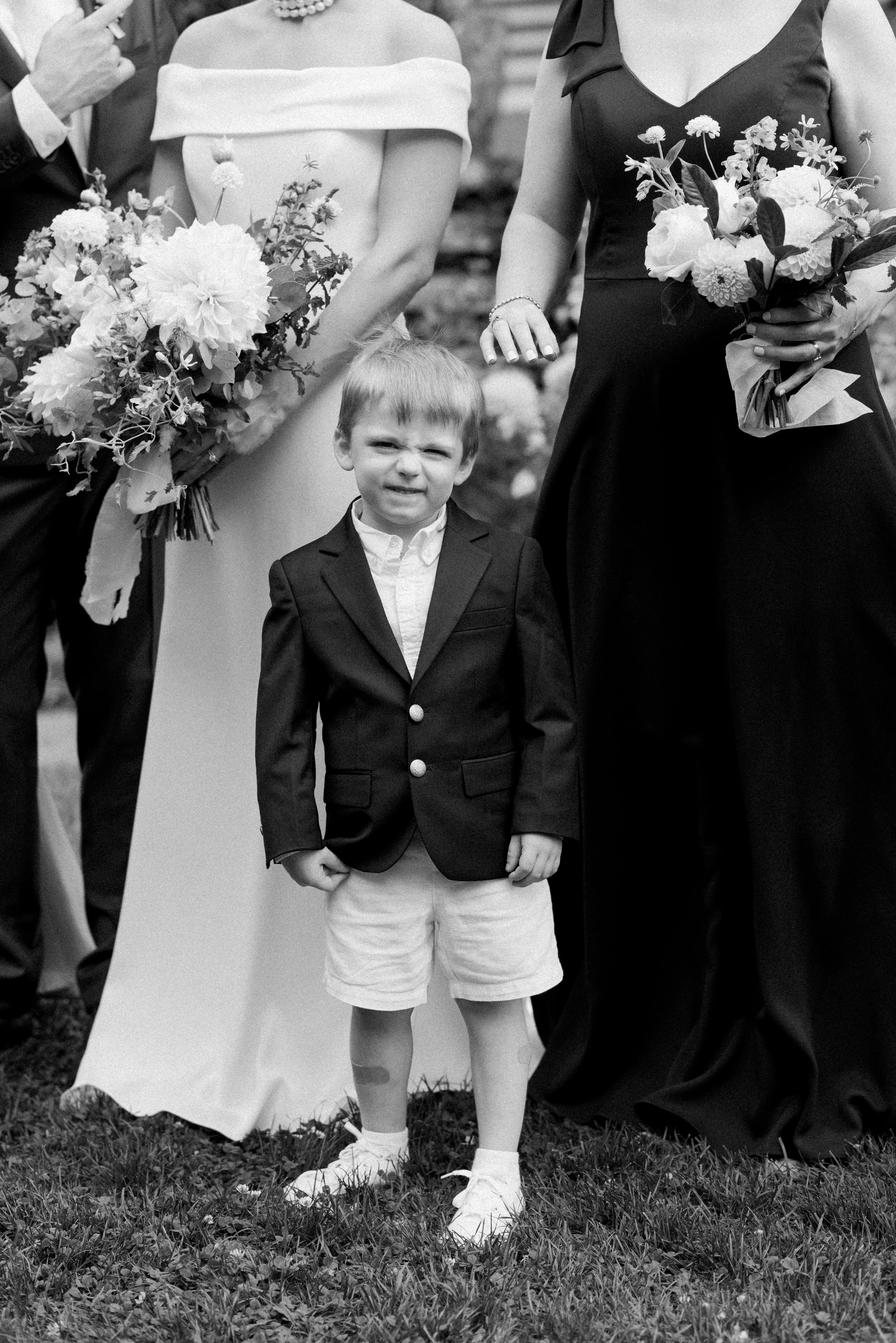 A young boy in a blazer, shorts, and sneakers standing in front of two women holding bouquets at a formal event, possibly a wedding.