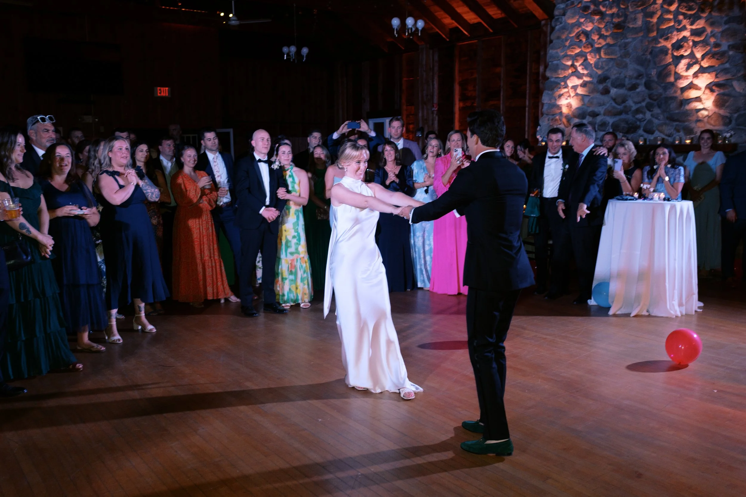 A bride and groom dancing at their wedding reception, surrounded by guests watching and taking photos in a rustic wooden venue.