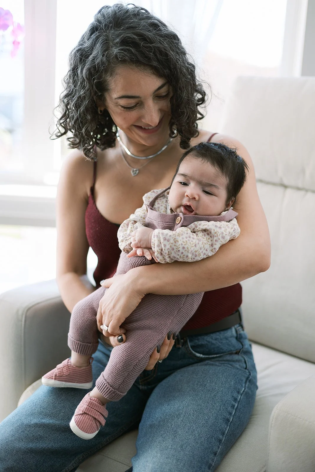 A woman with curly dark hair holding a young toddler girl sitting on her lap, smiling and looking down at the child in a cozy indoor setting with light-colored furniture and a bright window in the background.