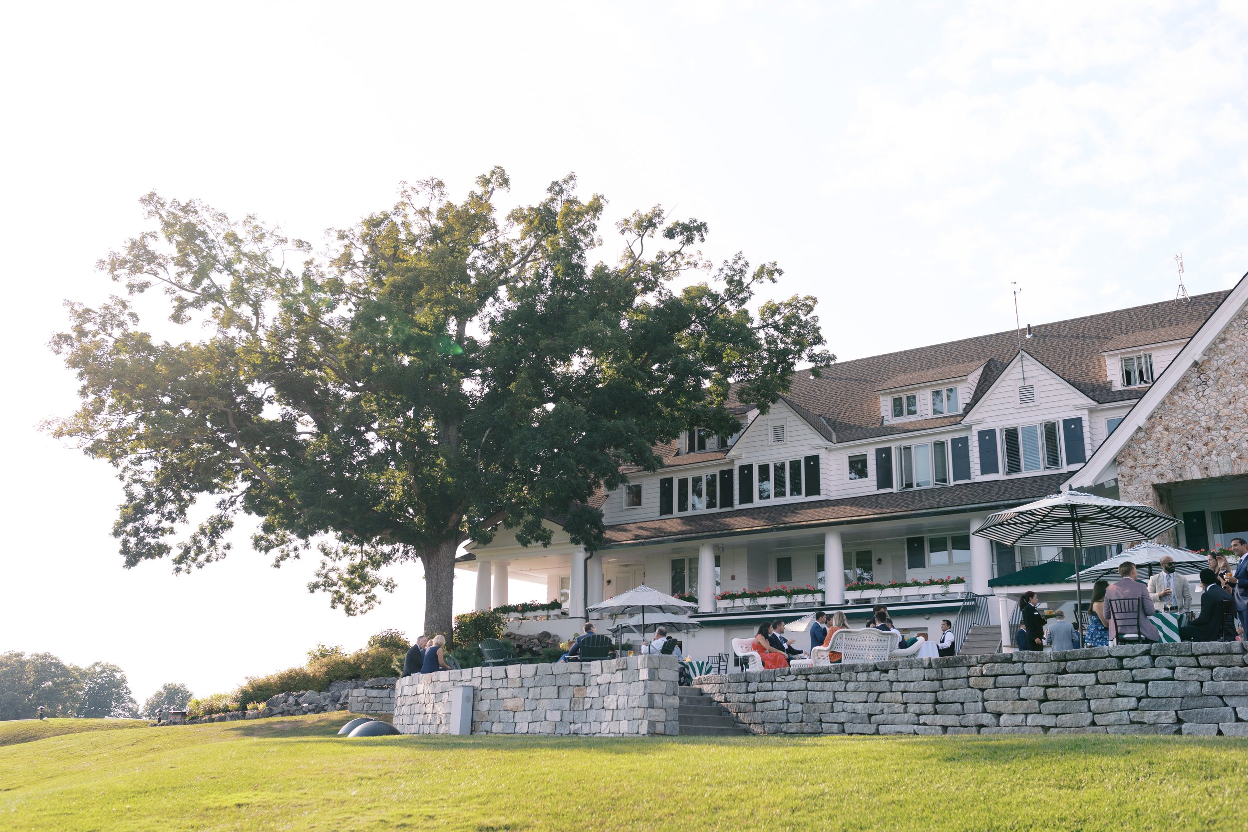 People enjoying outdoor gathering on a lawn with a large house and a big tree in the background in daytime.