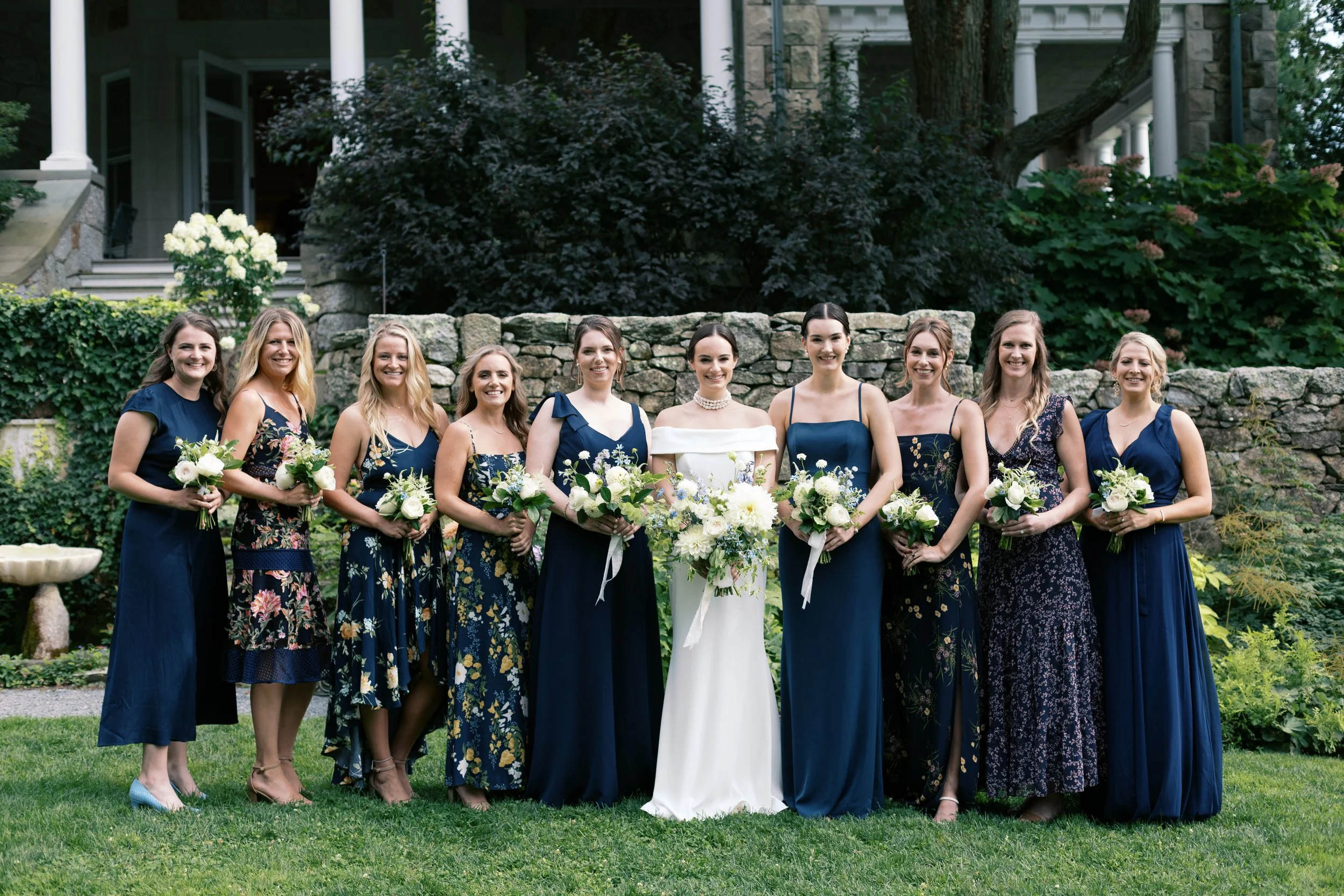 A bride and nine bridesmaids standing outdoors on a lawn, dressed in blue and floral dresses, holding bouquets of white and green flowers, in front of a stone wall and lush greenery.