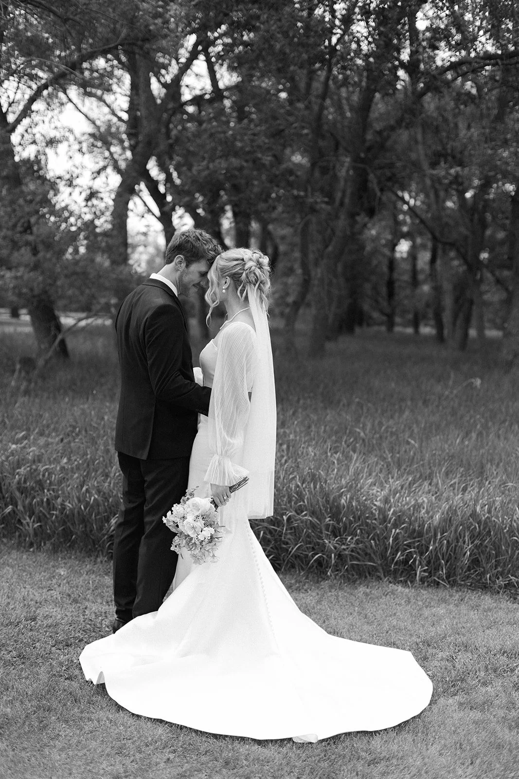 Black and white photo of a bride and groom standing close together outdoors, holding hands, with their foreheads touching, surrounded by trees.