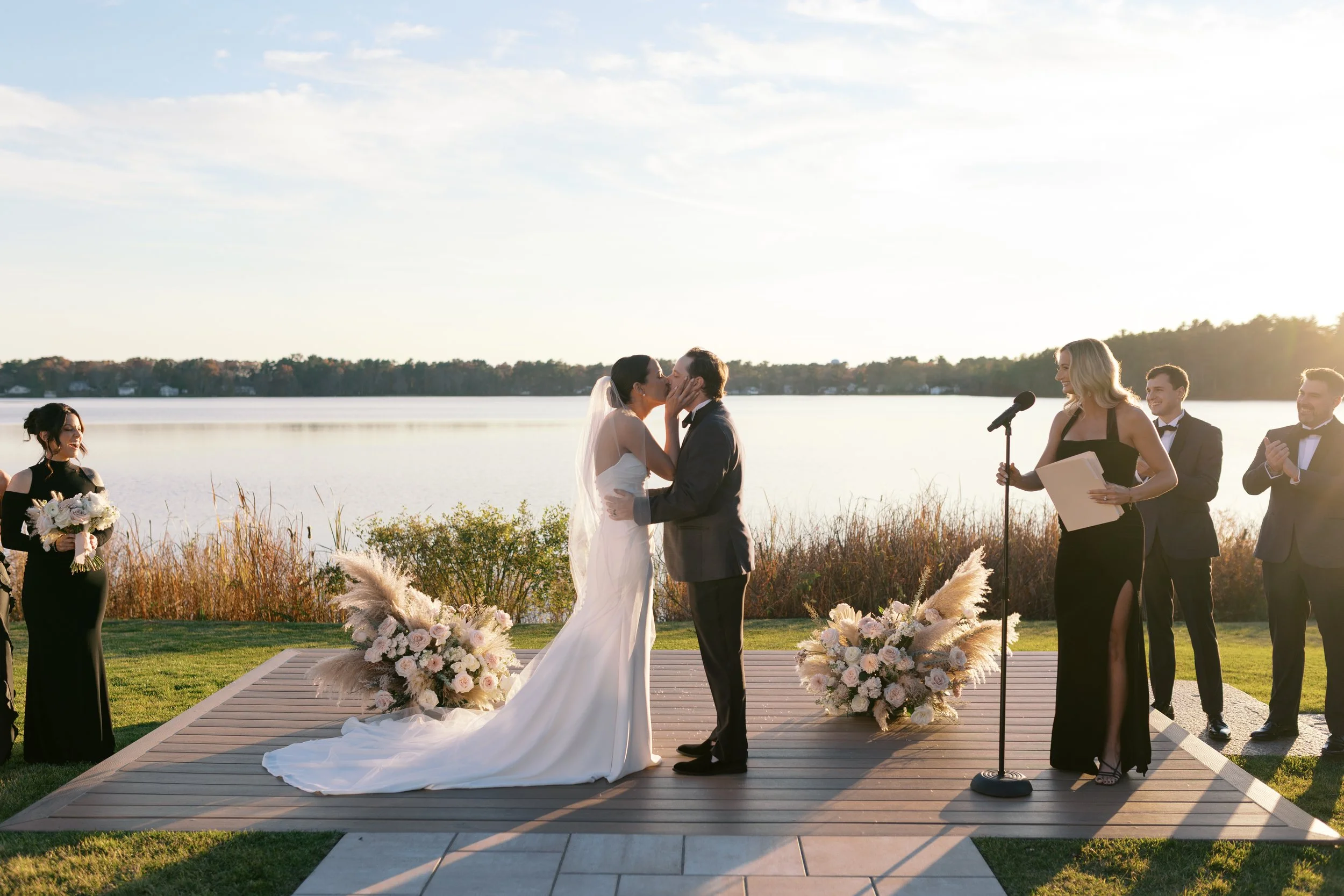 A wedding ceremony outdoors by a lake during sunset with the bride and groom kissing, officiant, bridesmaid, and groomsmen present.
