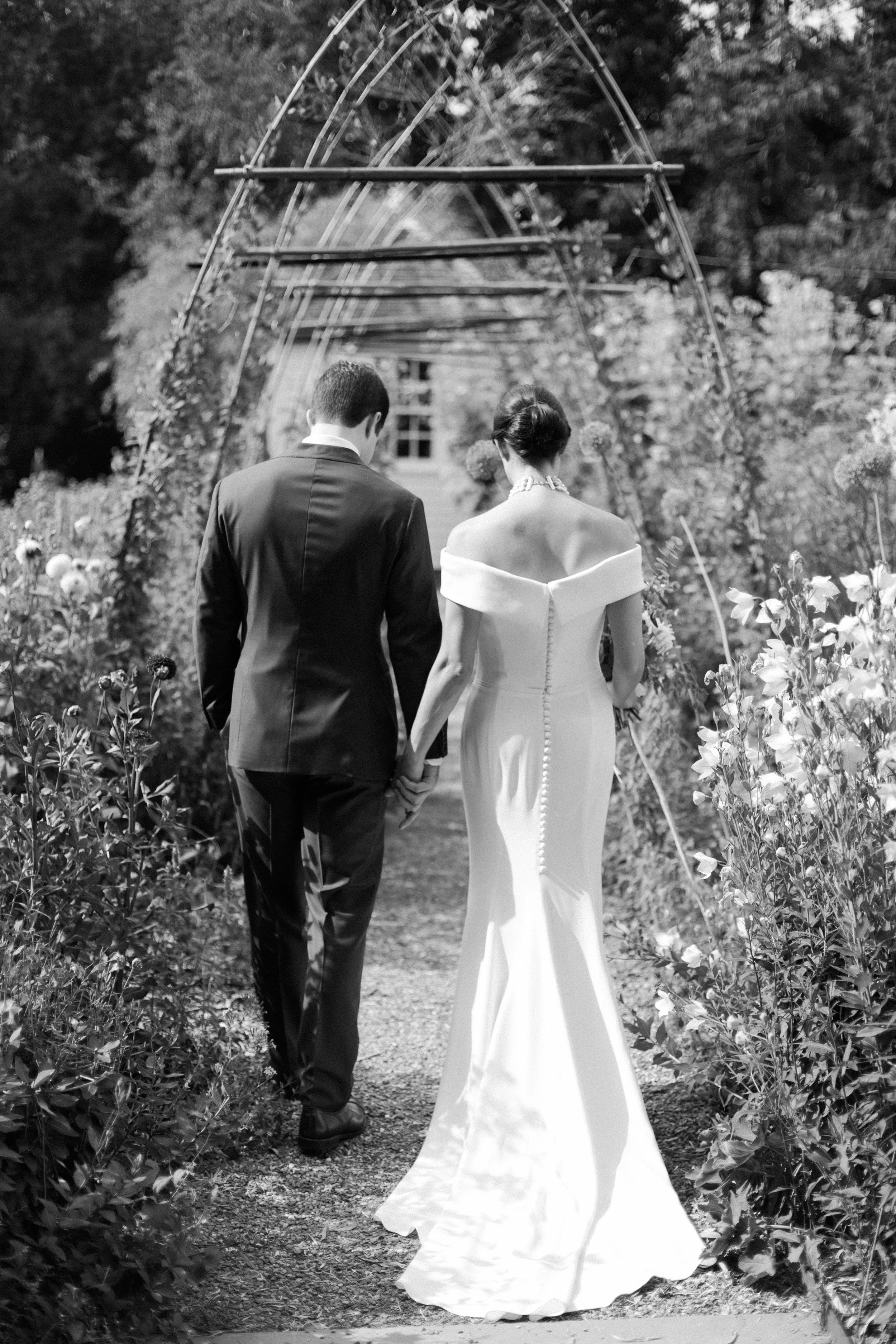 A bride and groom walking hand in hand through a garden archway, dressed in wedding attire; the woman wears a strapless gown and the man wears a suit.
