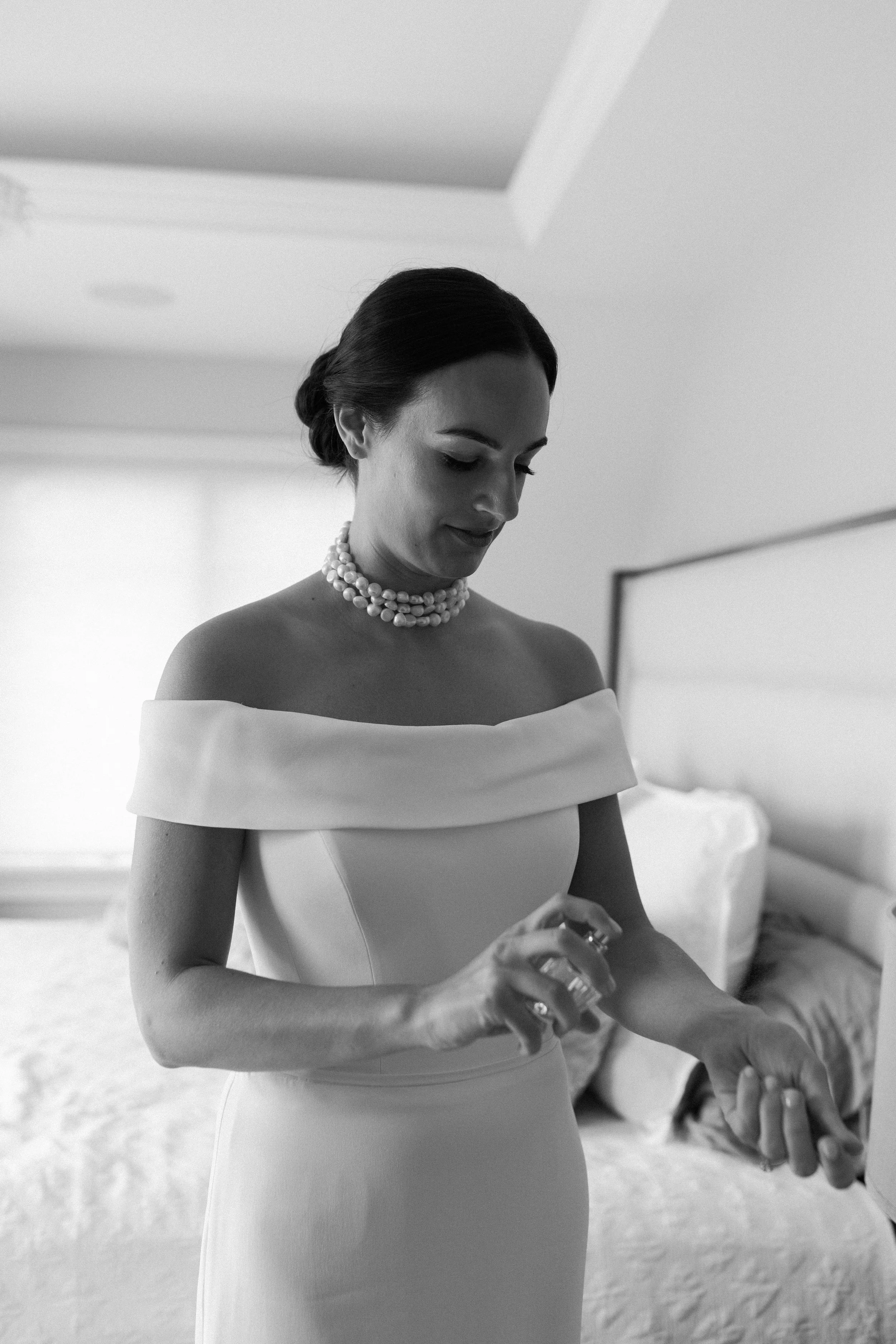 Black and white photo of a woman in an off-the-shoulder dress with pearl necklace, applying perfume in a bedroom.