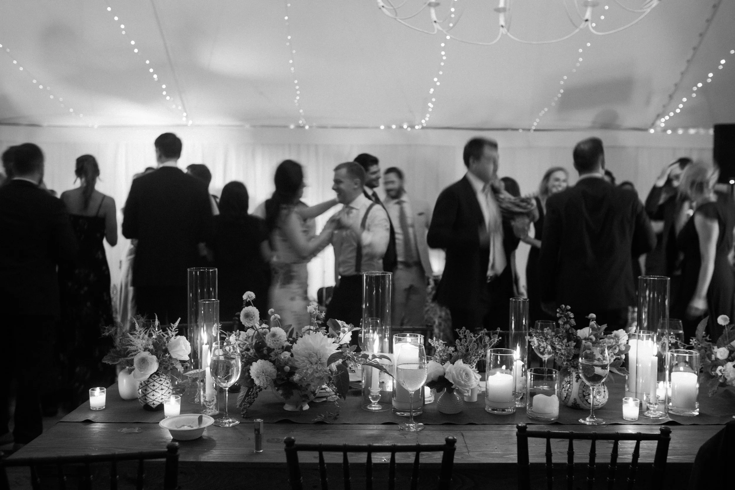 People dancing at a wedding or celebration with decorated table with candles and flowers in the foreground.