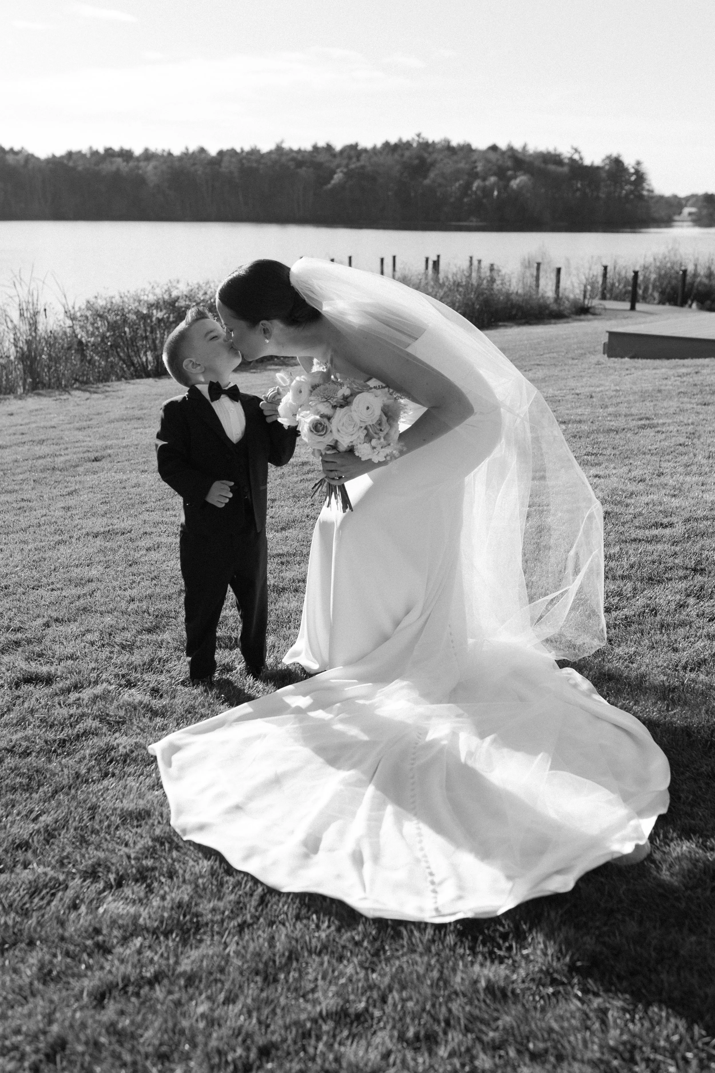 A bride in a wedding gown and veil kisses a young boy in a tuxedo by a body of water at sunset.