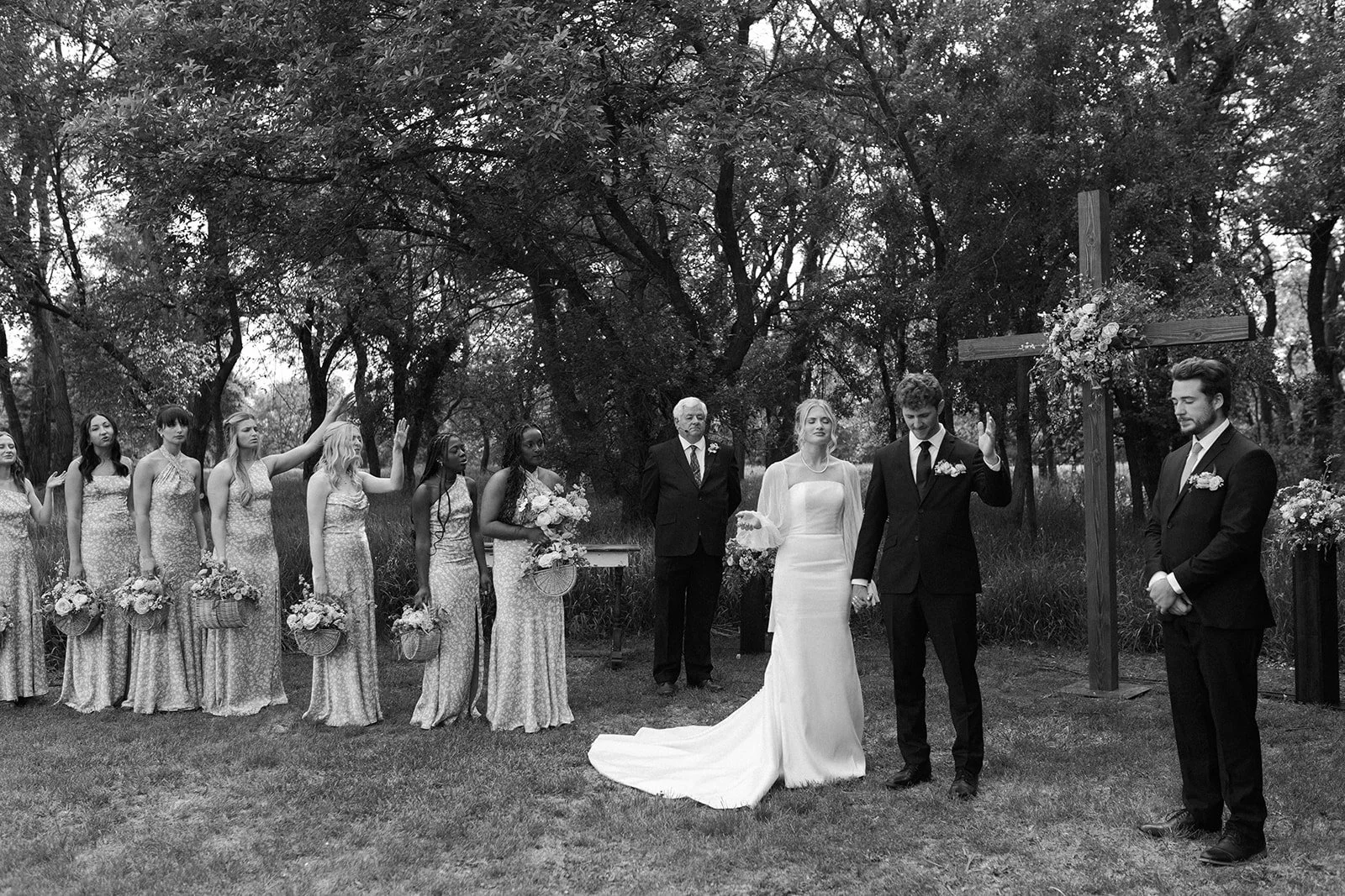 A black and white photo of a wedding ceremony outdoors in a wooded area. The bride and groom are holding hands in front of a large cross decorated with flowers. Bridesmaids and groomsmen, dressed in coordinated attire, stand on either side of the cou