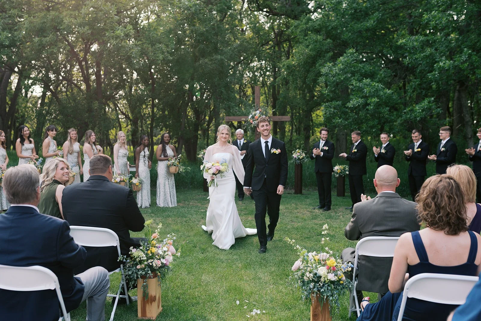 Bride and groom walking hand in hand down the aisle during outdoor wedding ceremony with guests seated on either side, bridesmaids and groomsmen standing behind, surrounded by trees and greenery.