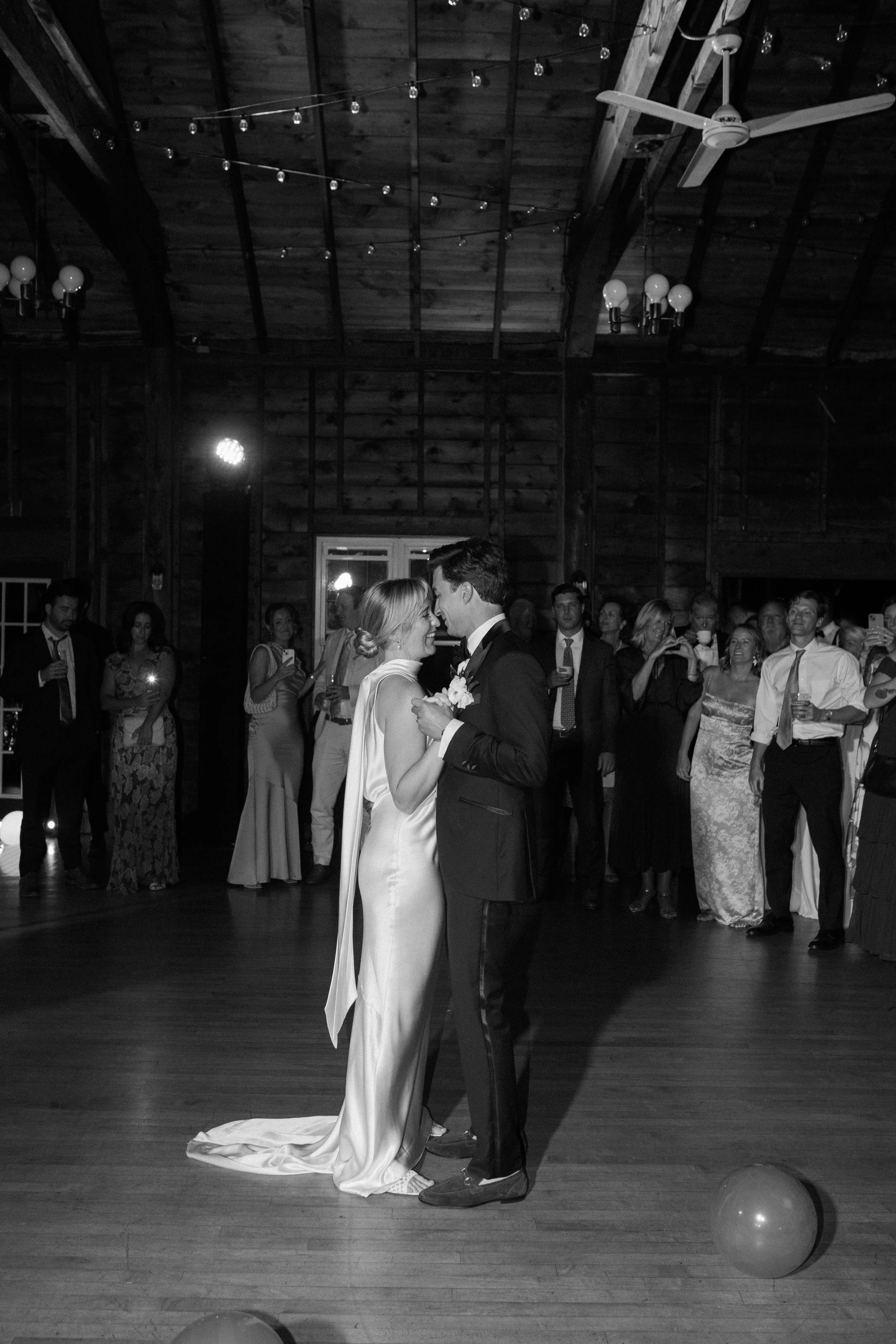 A bride and groom sharing a dance at their wedding reception in a rustic hall with wooden walls, surrounded by guests.