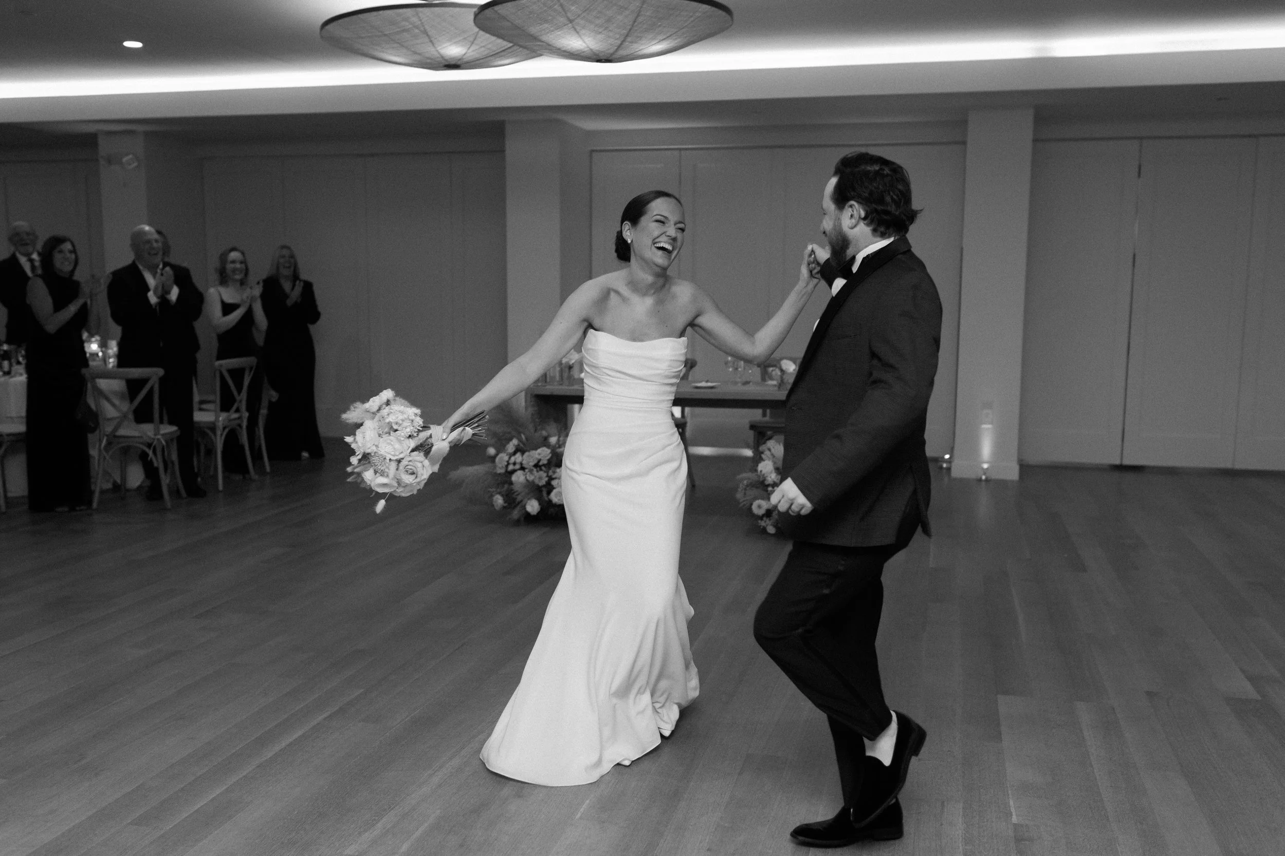 A bride and groom dancing and laughing at their wedding reception, surrounded by seated guests clapping, in a decorated indoor venue.