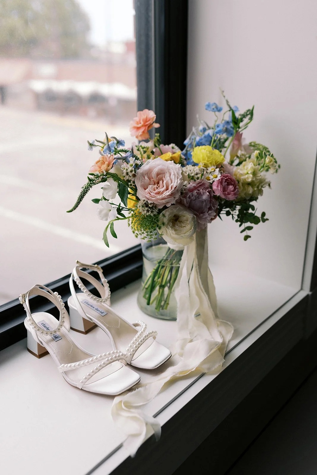 A pair of white high-heeled sandals with pearl embellishments on the straps, placed on a windowsill next to a glass vase of colorful flowers.