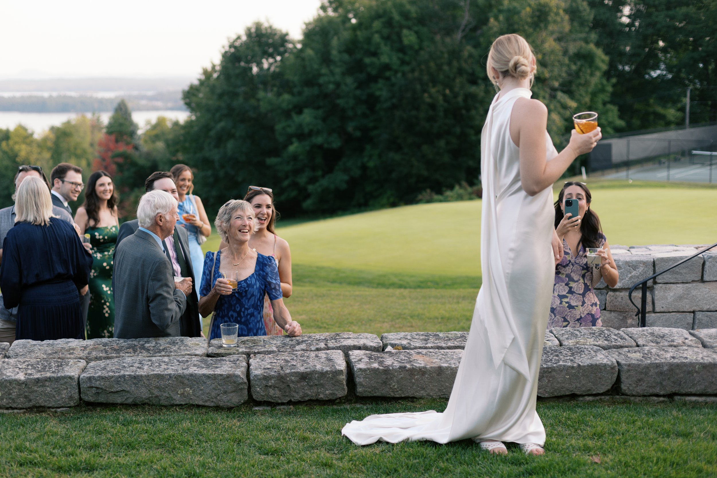 A woman in a white dress holding a drink stands in front of a group of people outdoors during a celebration. The group, including elderly and younger adults, appears to be enjoying themselves and smiling. The setting is a lush, green area near a tenn