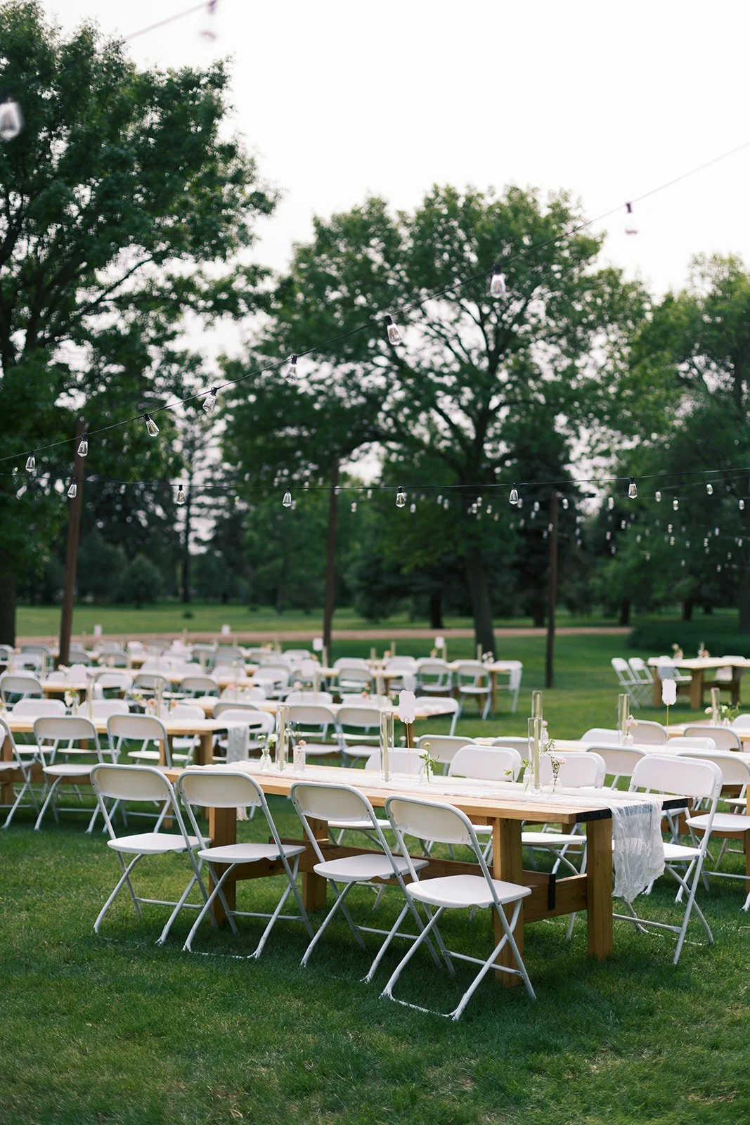 Outdoor event setup with tables and white chairs under string lights in a park.
