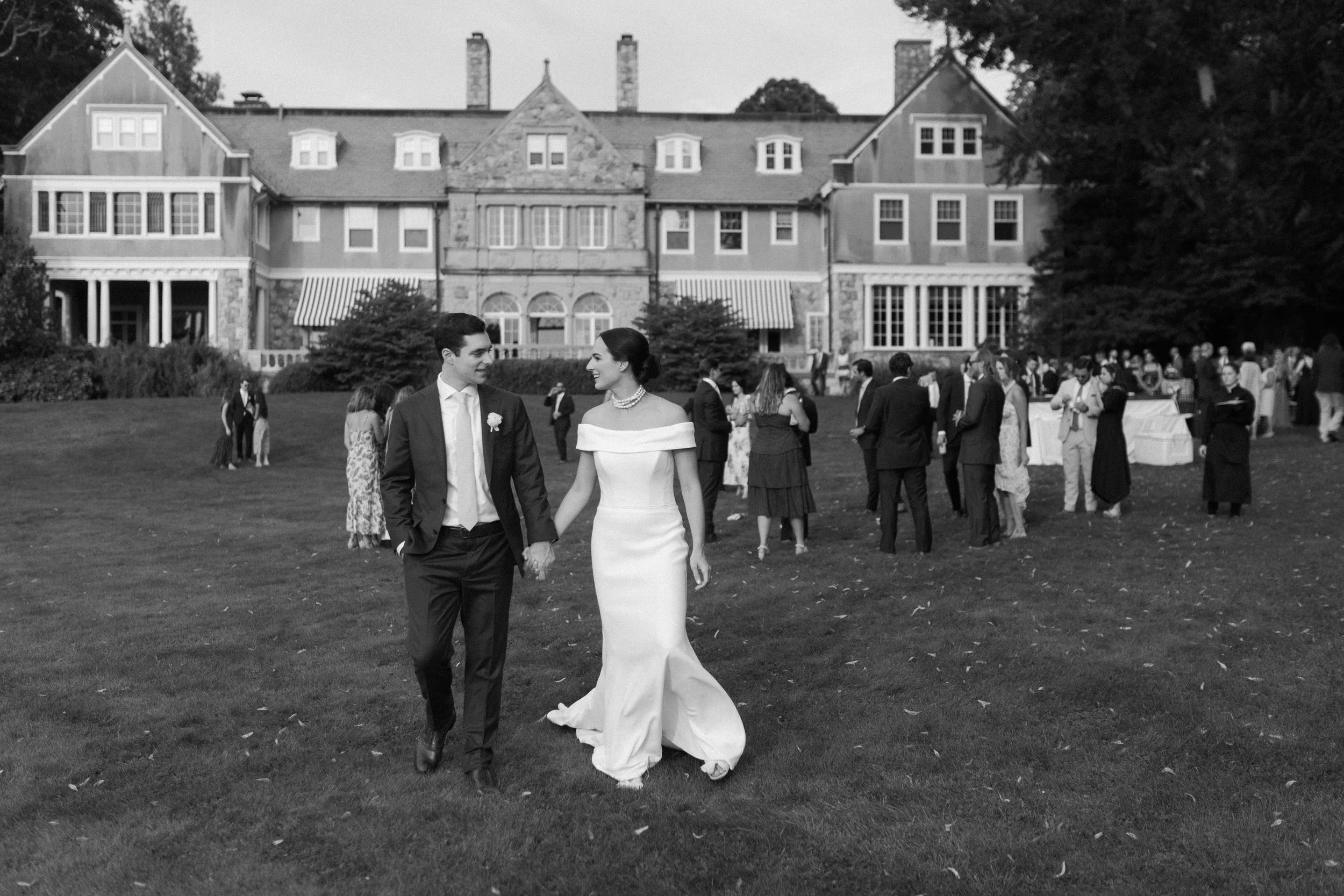 Black and white photo of a bride and groom walking hand-in-hand on a grassy lawn outside a large, historic house with multiple stories and a stone facade. The bride is wearing an off-the-shoulder wedding dress and the groom is dressed in a suit. In t