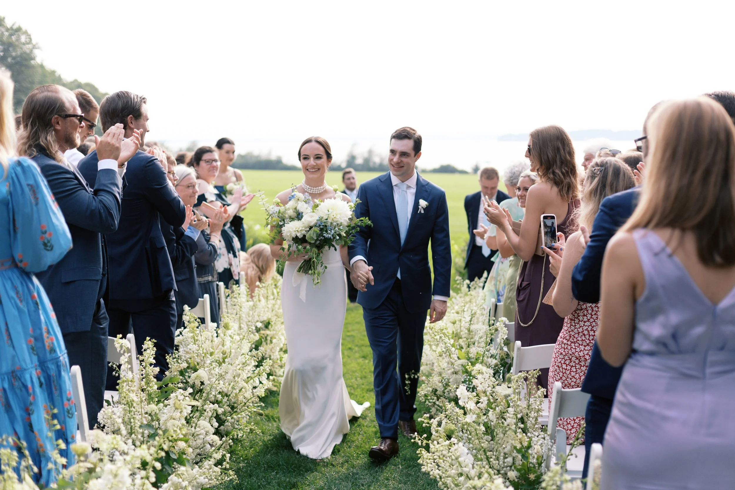 Bride and groom walking down the aisle after their wedding ceremony outdoors, surrounded by friends and family, on a green lawn with flowers lining the aisle.