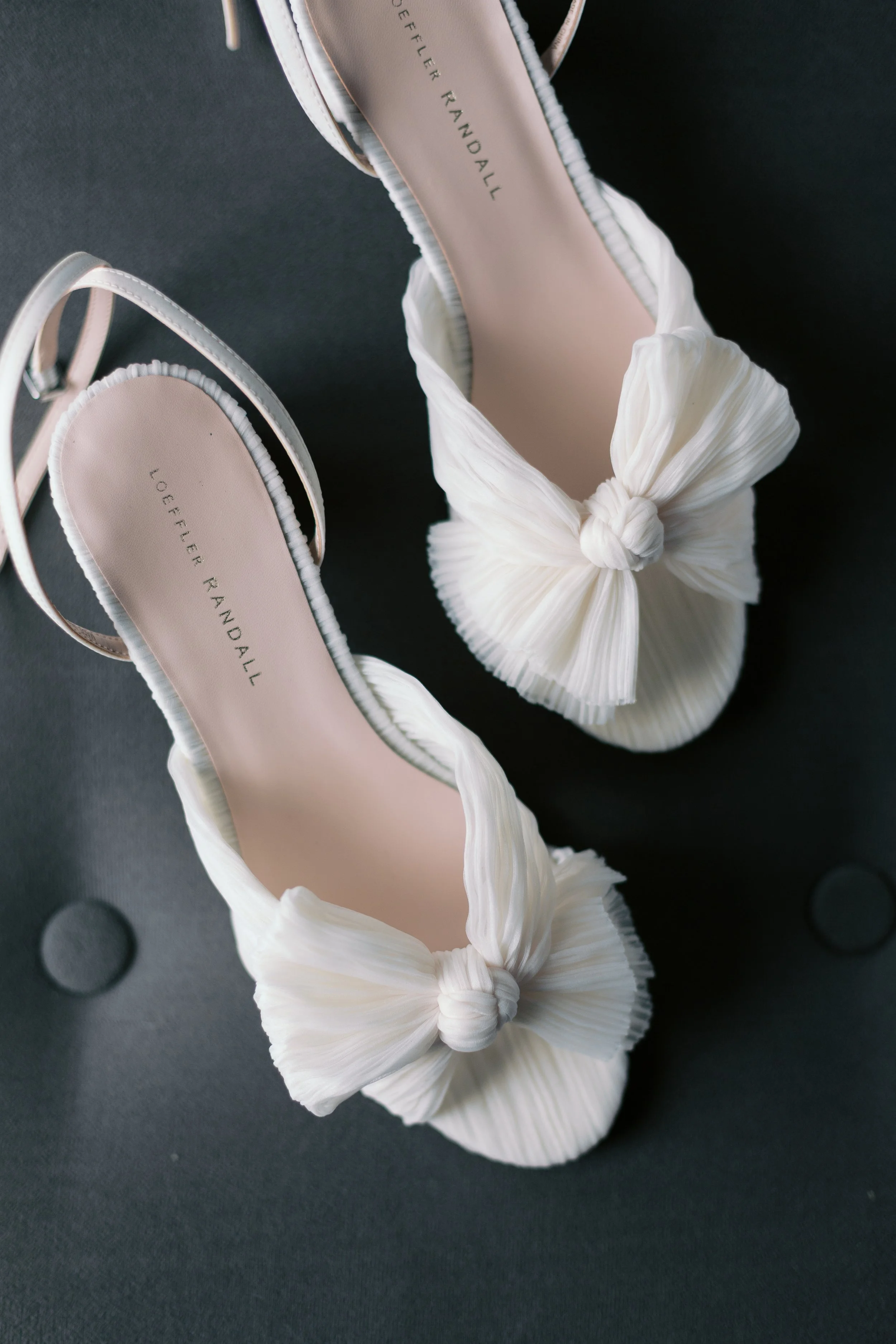 Pair of white pleated high-heeled shoes with bow detail, placed on a black surface.