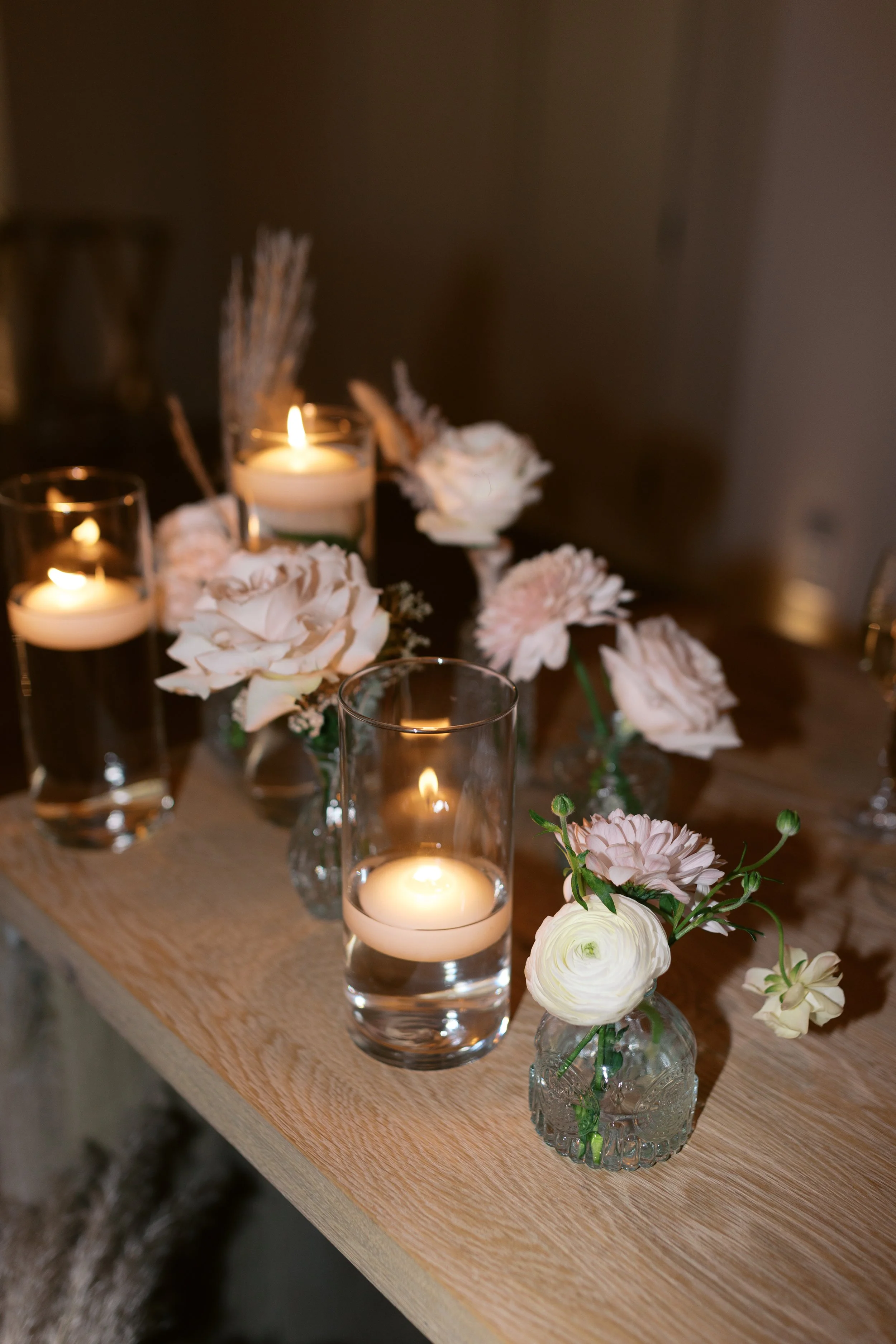 Vases with pink and white flowers and candles in glass holders on a wooden table.