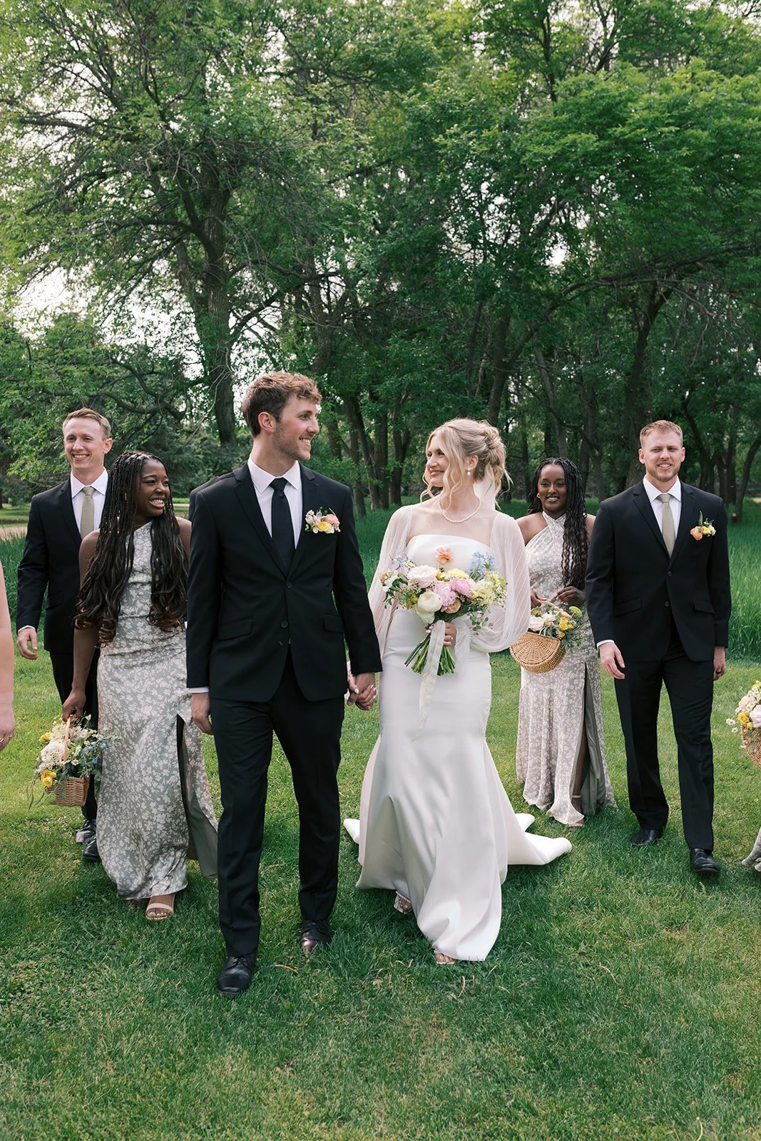 A bride and groom holding hands walking outdoors with friends and bridesmaids in a lush green park.