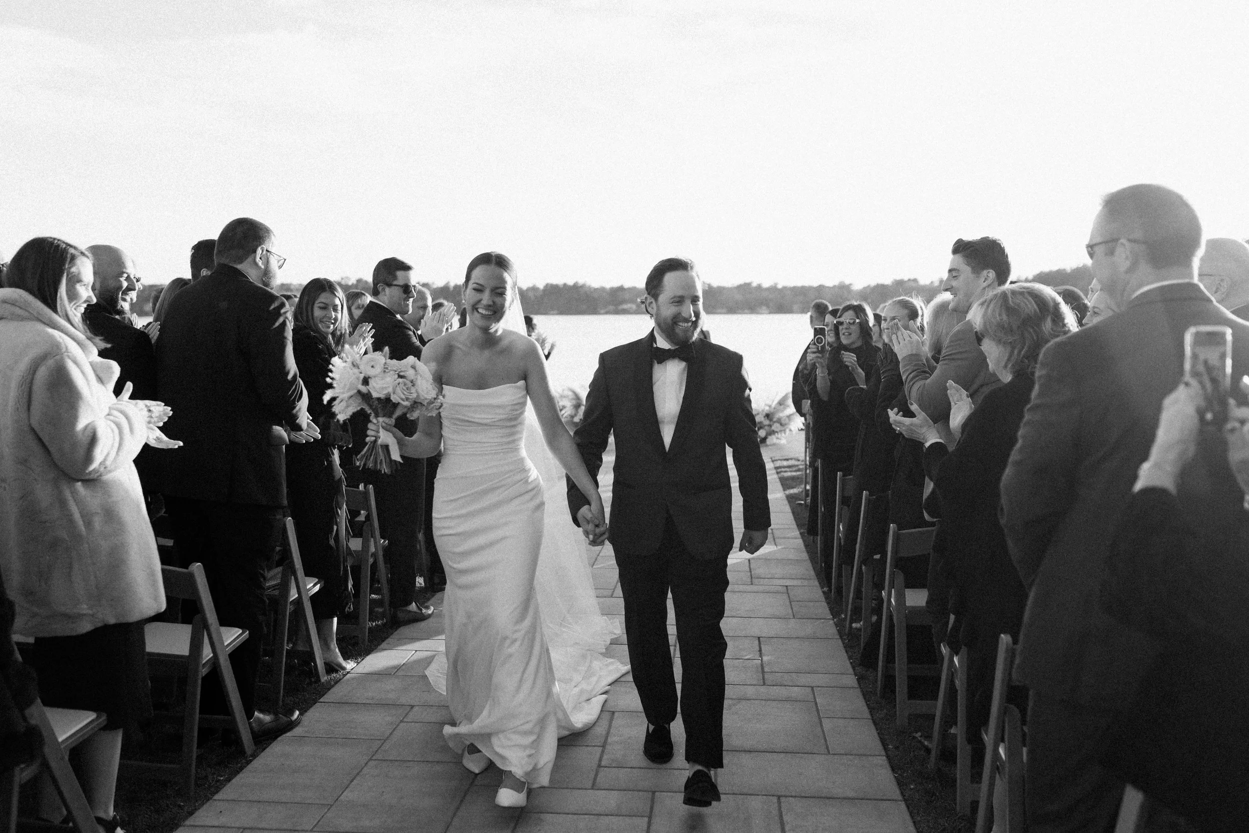 A black-and-white photo of a bride and groom walking hand in hand down an outdoor aisle, smiling, surrounded by seated and standing guests during a wedding ceremony near a body of water.