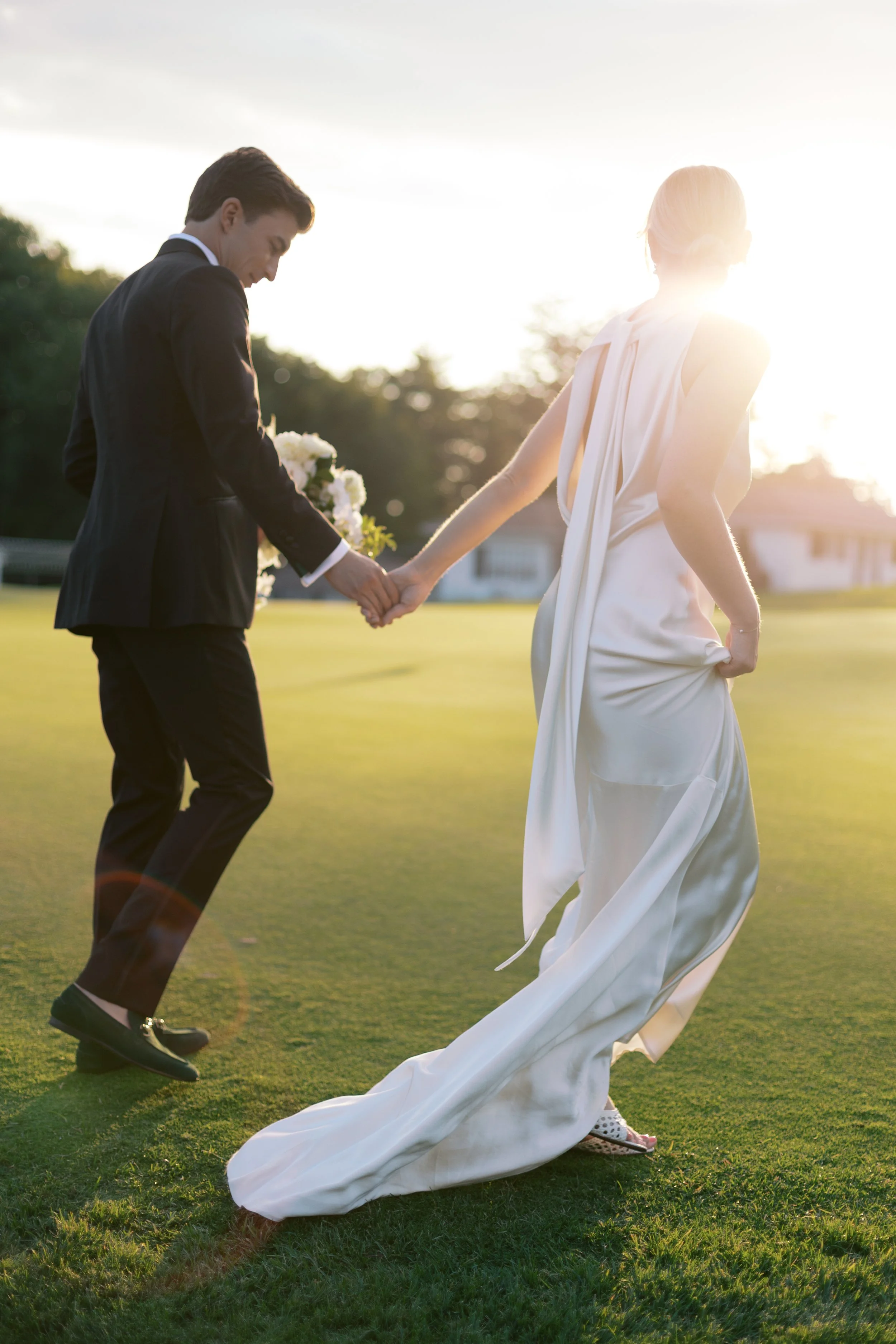 A newlywed couple holding hands and walking on a grassy field at sunset, with the bride's long white dress flowing and the groom in a black suit holding a bouquet of white flowers.