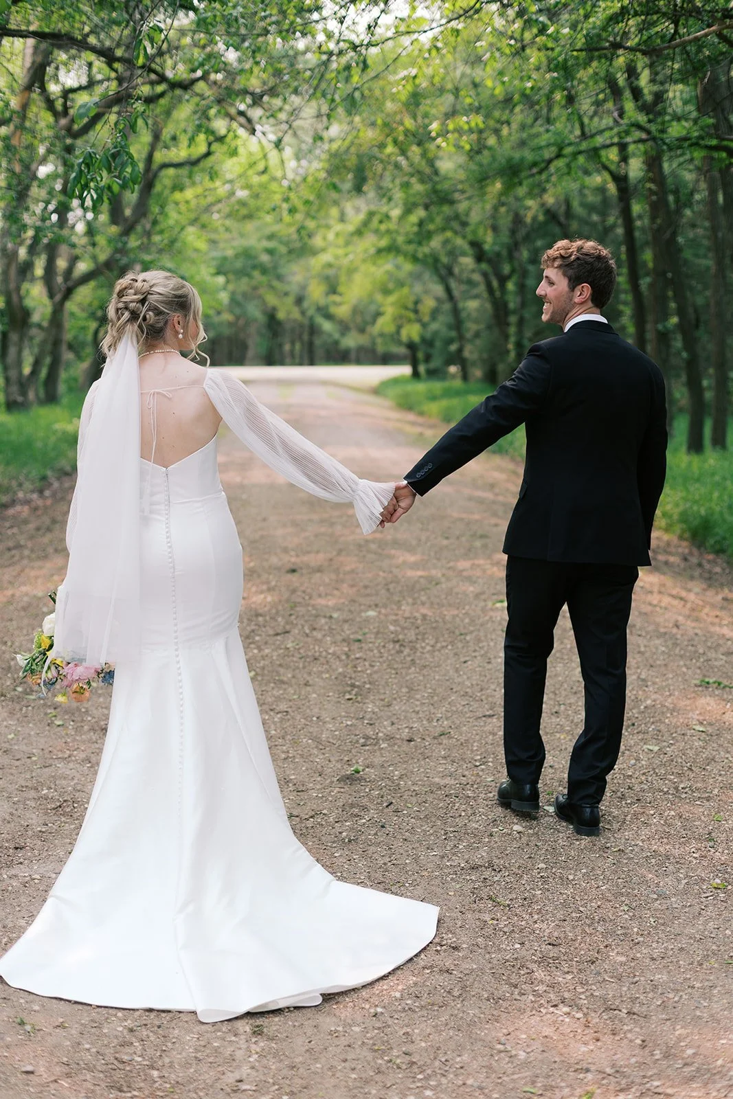 A bride and groom holding hands and walking in a wooded outdoor setting.