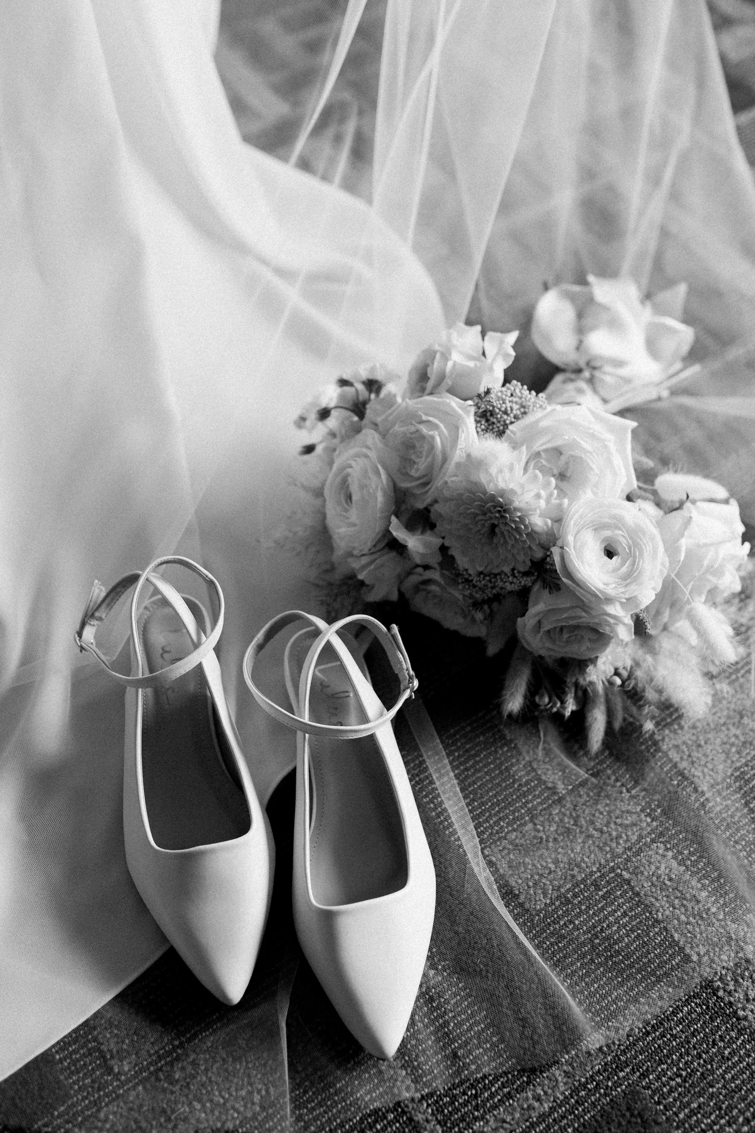 A pair of white high-heeled shoes with ankle straps, a bridal bouquet with roses and other flowers, and a veil on a textured surface.