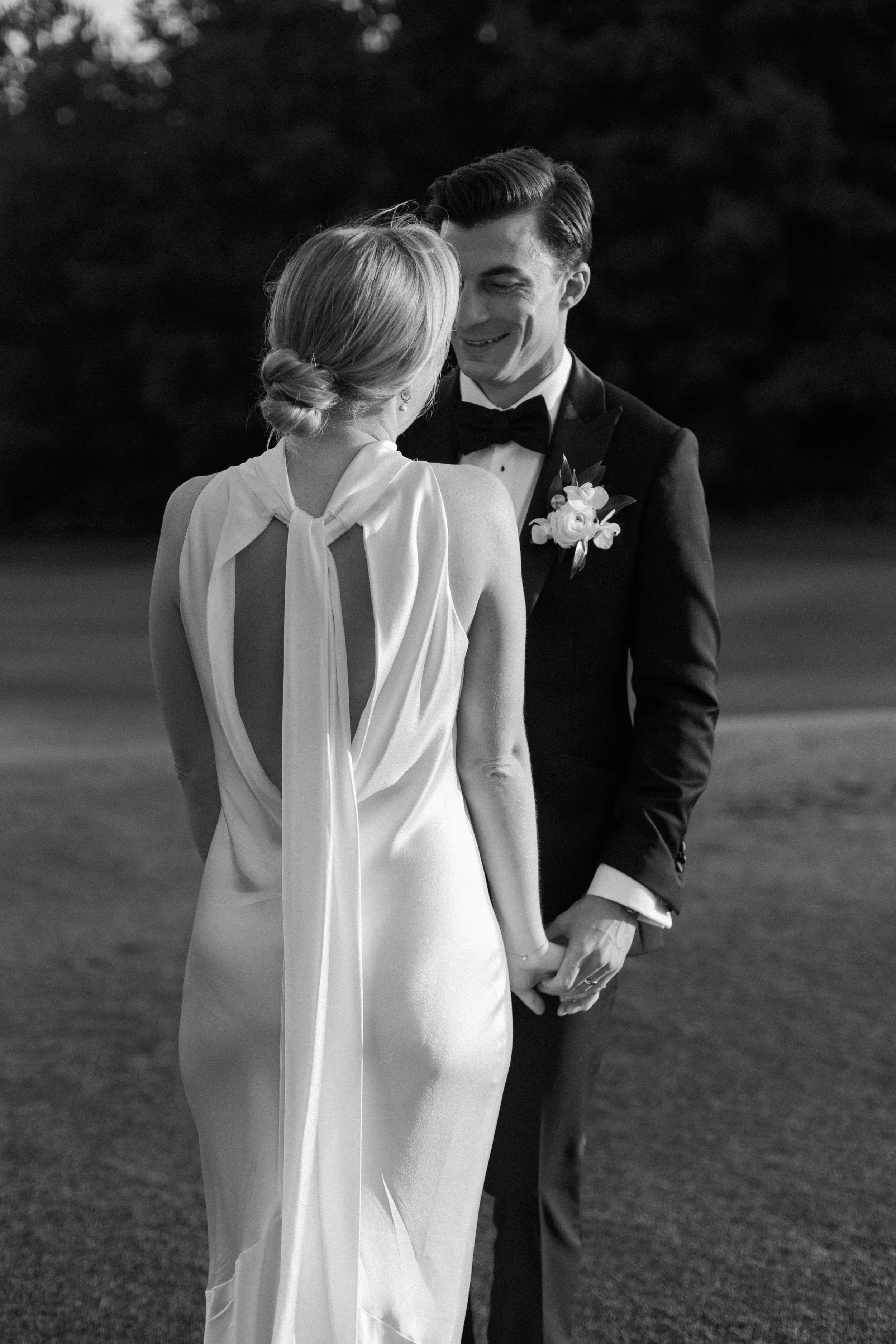 A bride and groom holding hands and smiling, standing outdoors during their wedding at dusk.