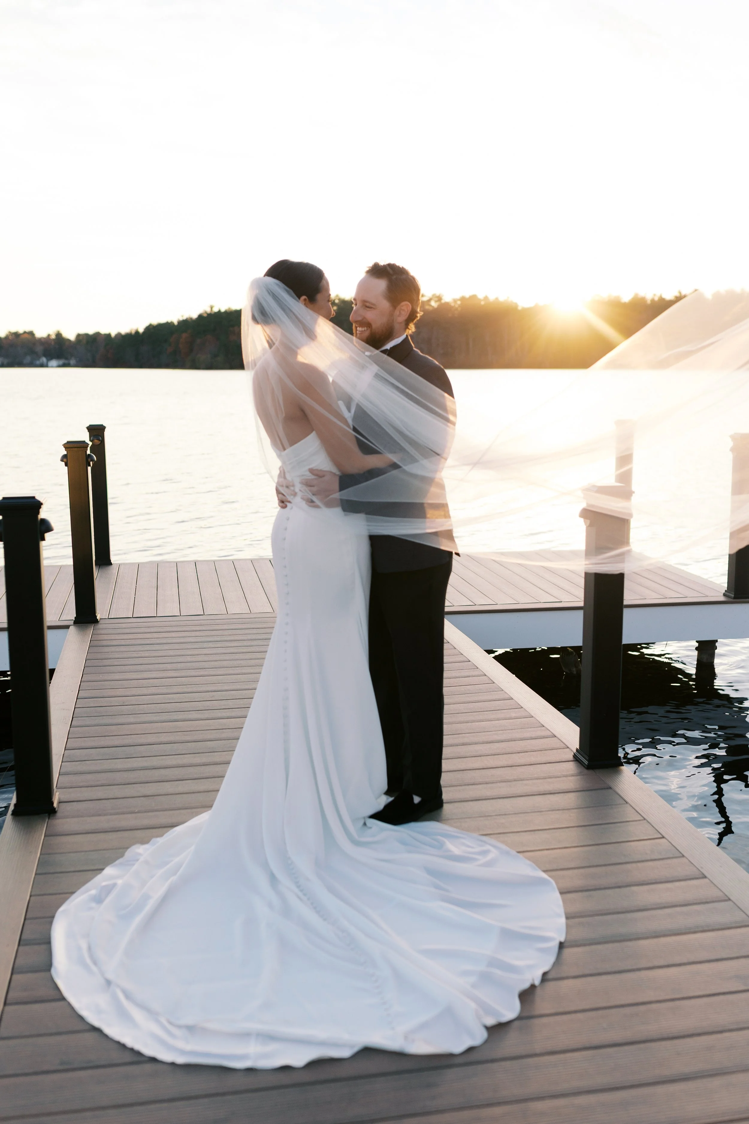 A bride and groom share a kiss on a dock by the water at sunset, with the bride in a white gown and veil, and the groom in a tuxedo.