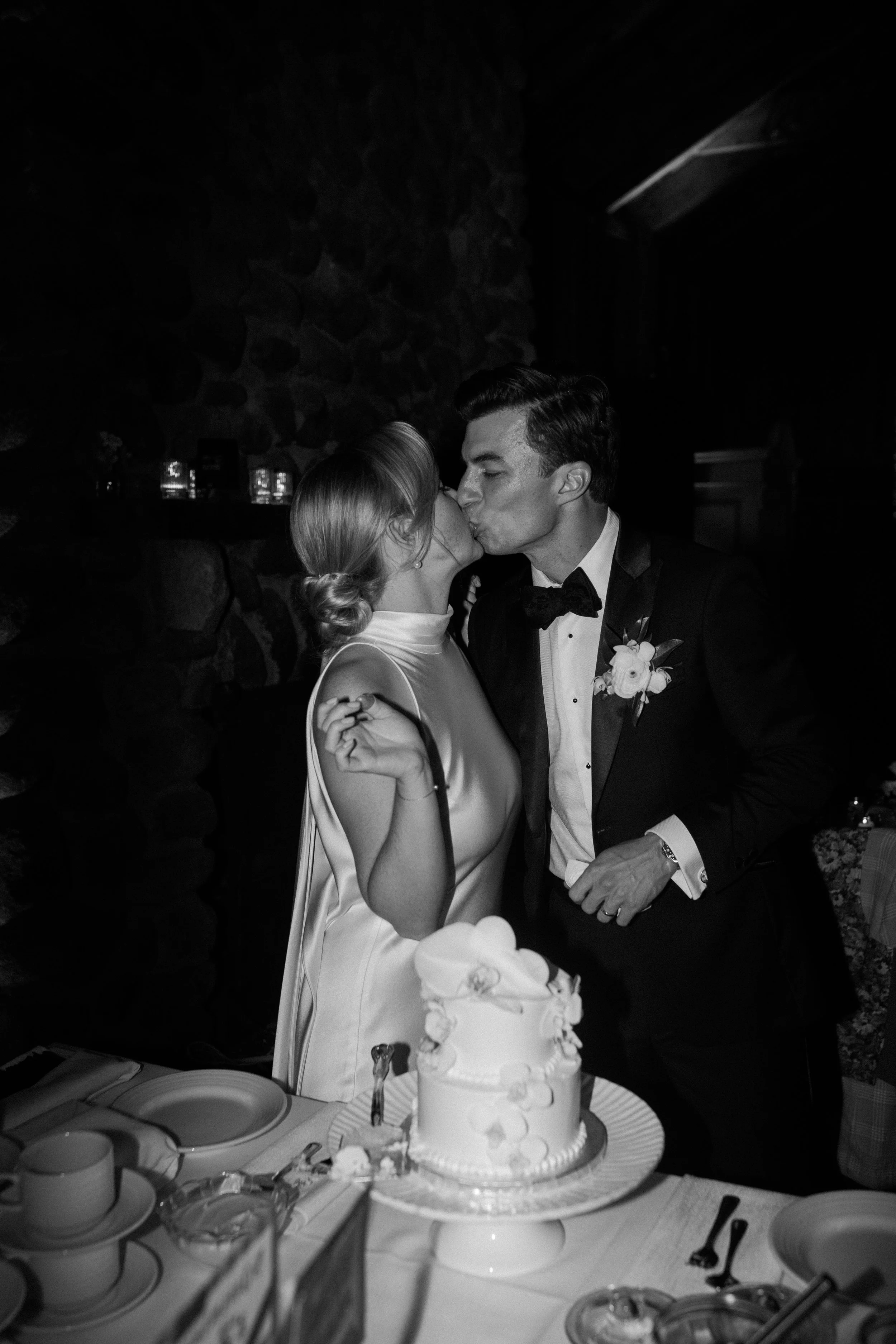Black-and-white photo of a bride and groom kissing at their wedding reception, with a wedding cake on the table in front of them.