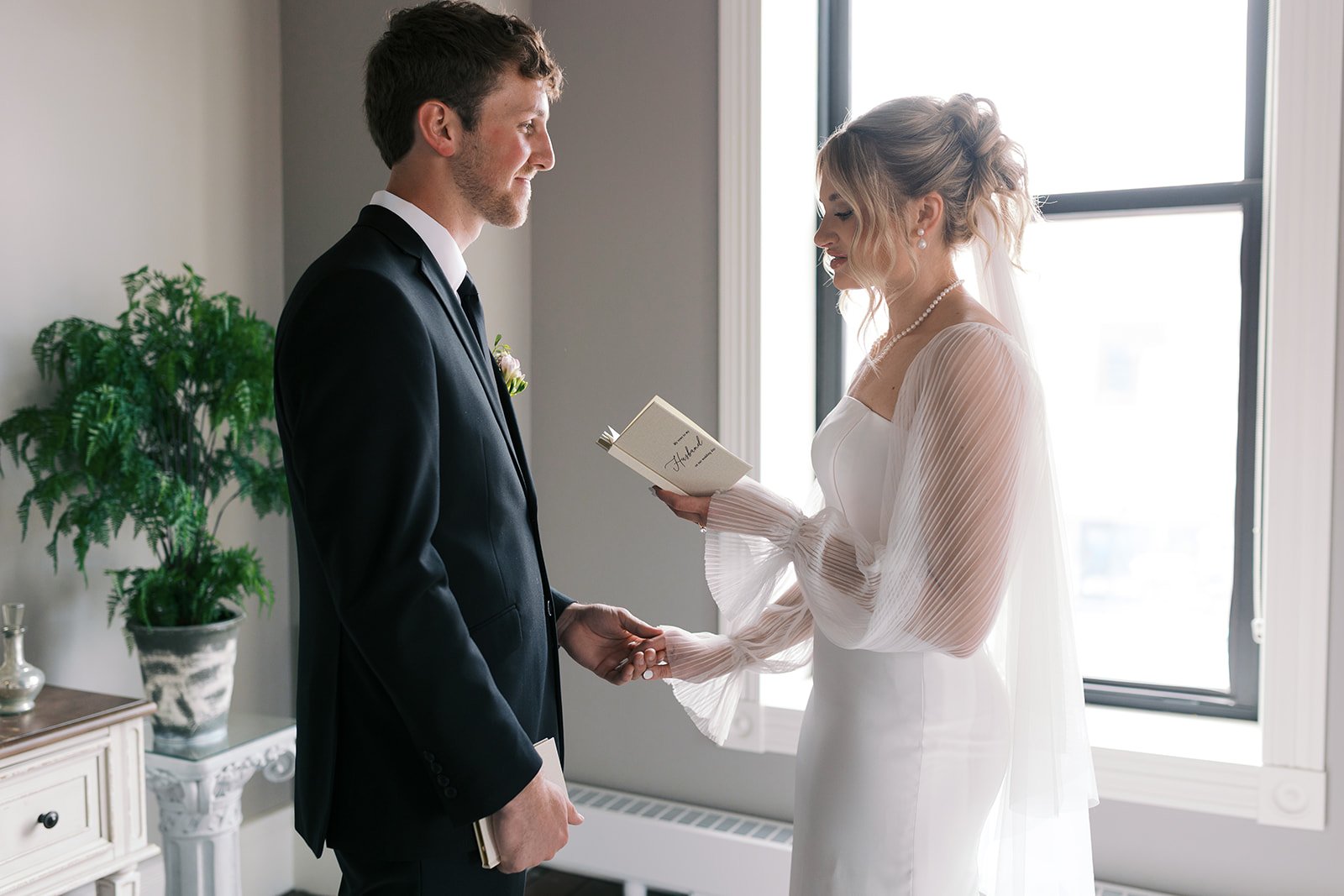 A bride and groom exchanging vows during a wedding ceremony indoors, holding hands and standing in front of a window with sunlight streaming in.