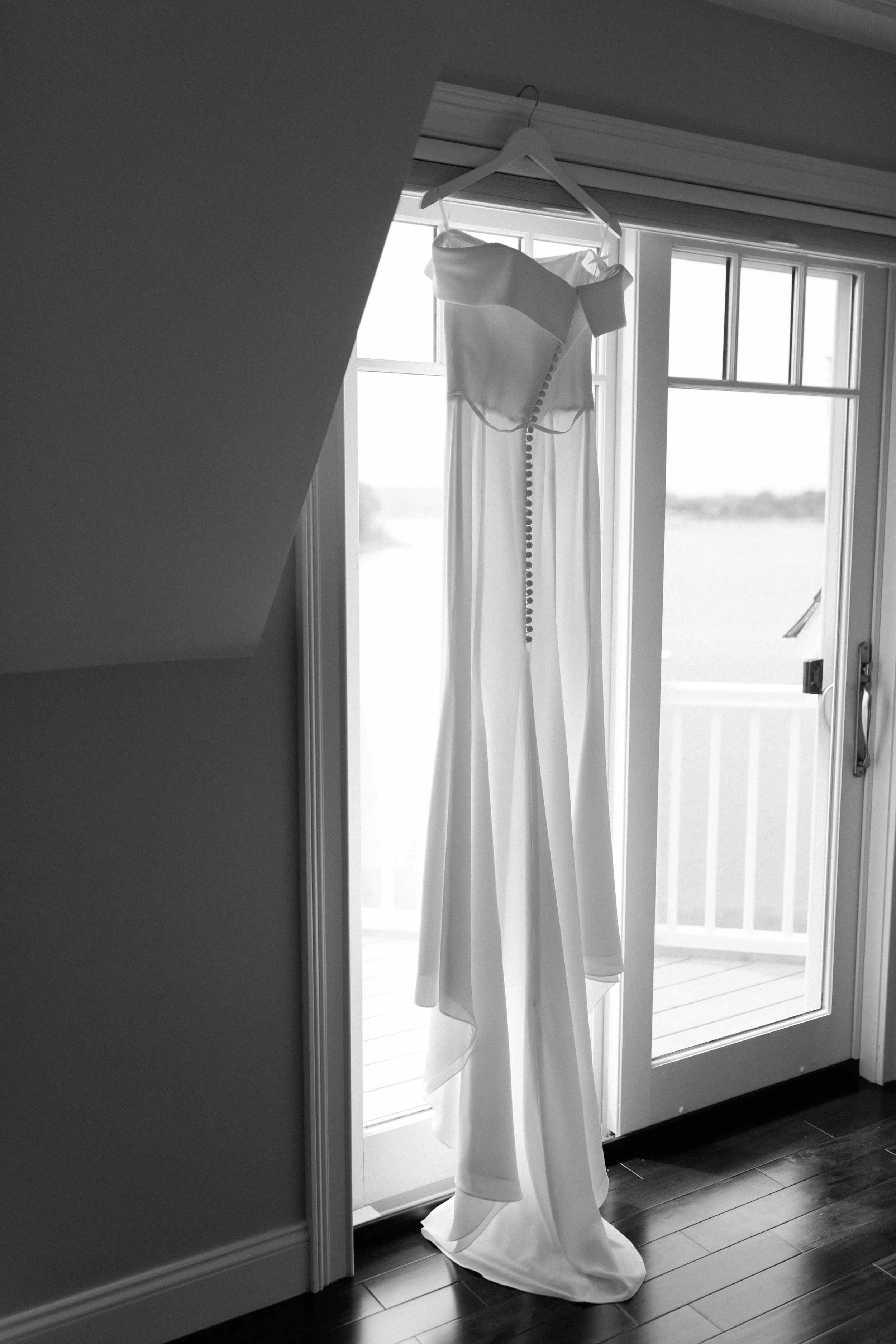 White wedding dress hanging on a hanger on a window frame, with a balcony door in the background.