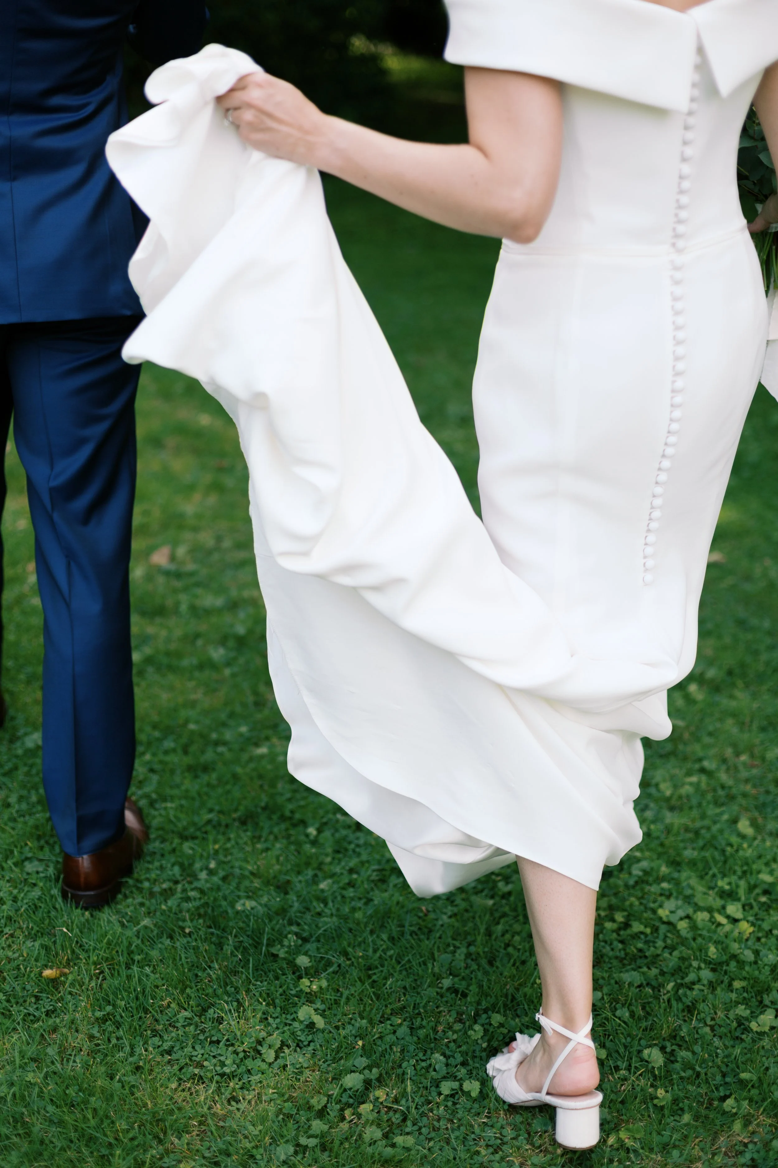 Bride in a white dress and heels adjusting her wedding gown outdoors, with a man in a blue suit nearby.