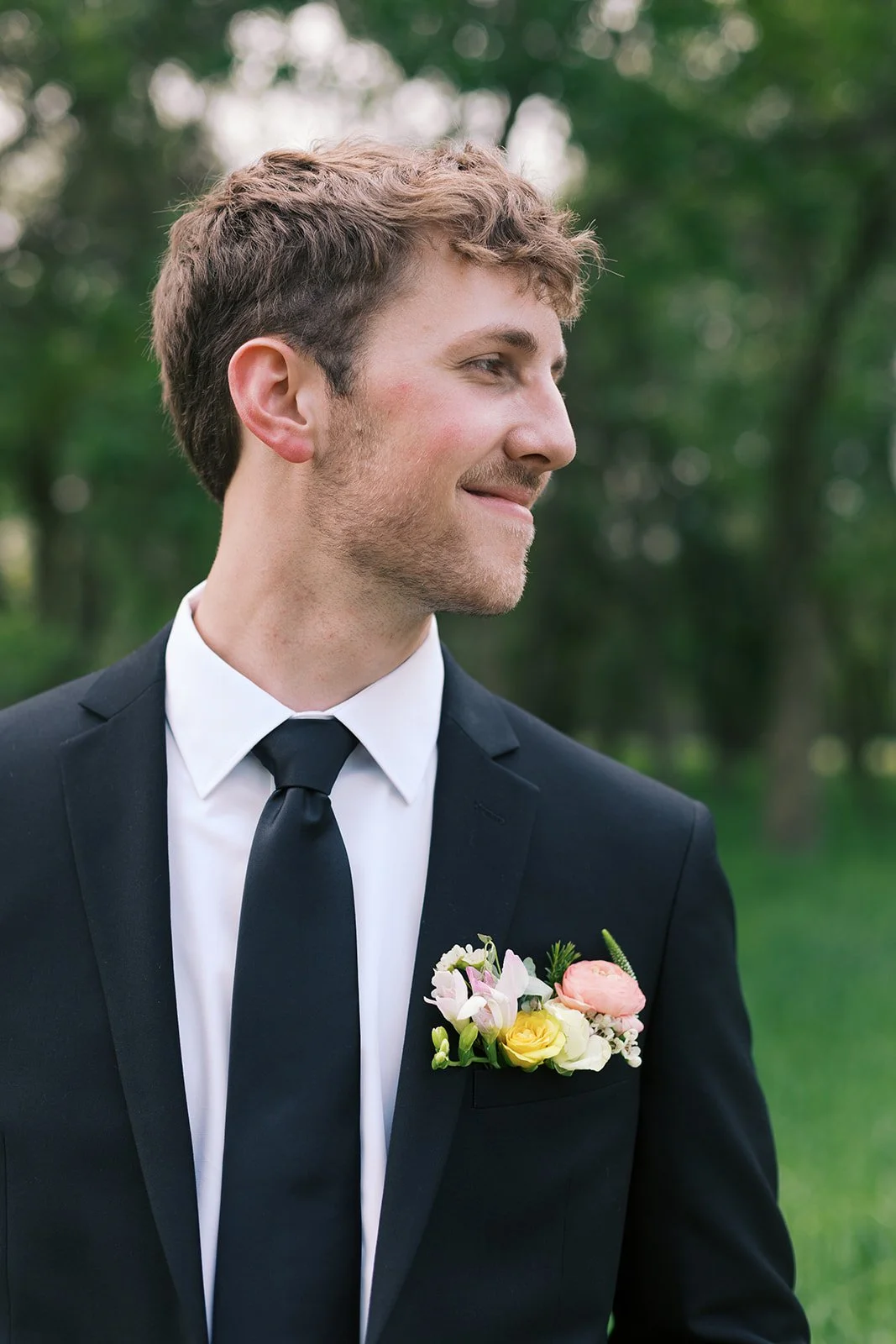 A young man in a black suit and tie with a white shirt, standing outdoors with trees in the background, smiling and looking to the side. He has a boutonniere with pink, white, and yellow flowers pinned to his suit lapel.