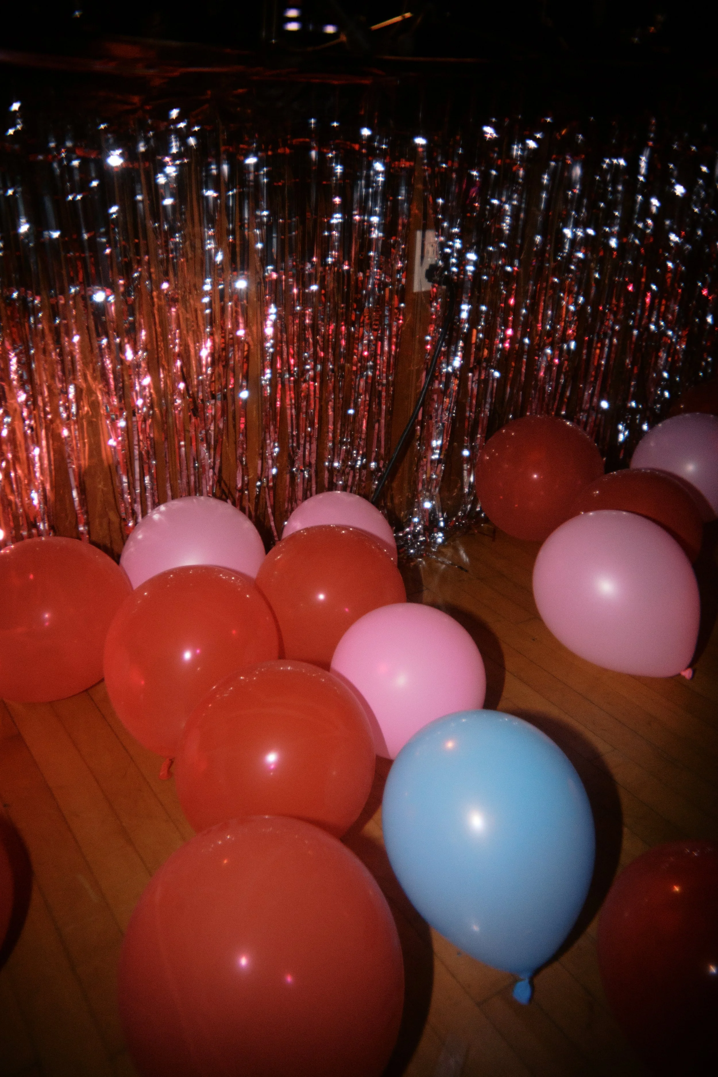 A floor decorated with colorful balloons including red, pink, and blue, against a backdrop of shimmering silver and red streamers, likely at a party or celebration.